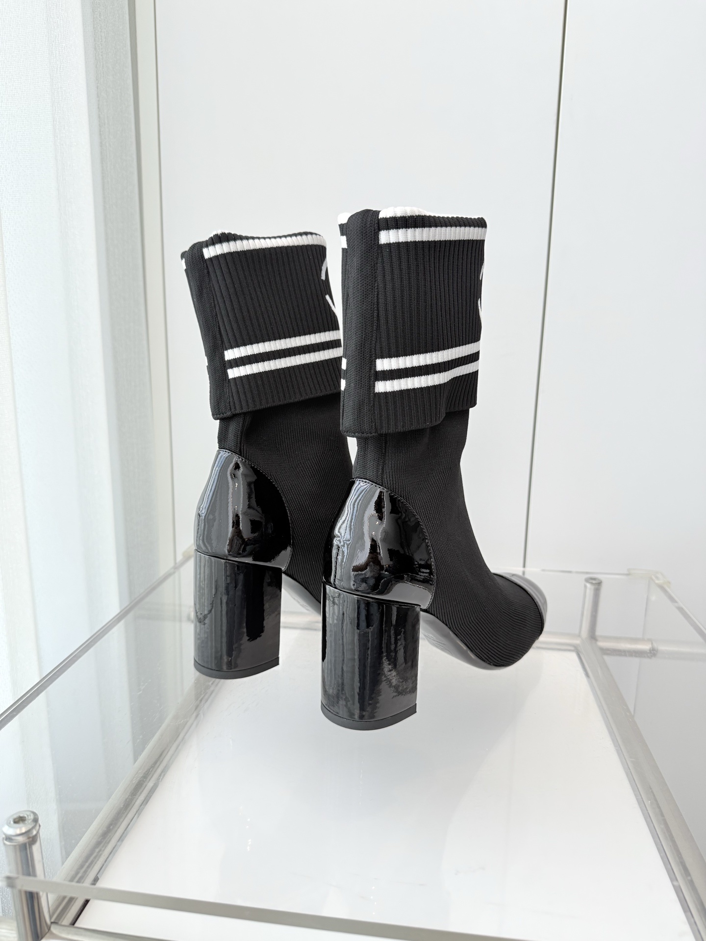 A pair of elegant black knit sock boots with patent leather cap toes and white logo fold-over cuffs displayed on a glass shelf. - Hình 7