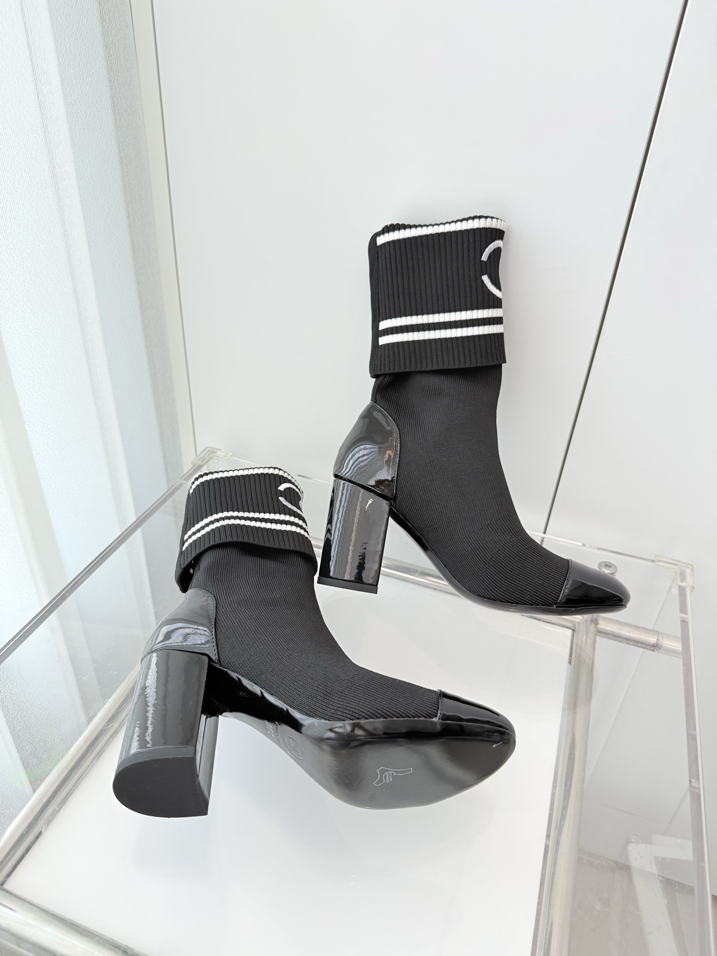A pair of elegant black knit sock boots with patent leather cap toes and white logo fold-over cuffs displayed on a glass shelf. - Hình 6