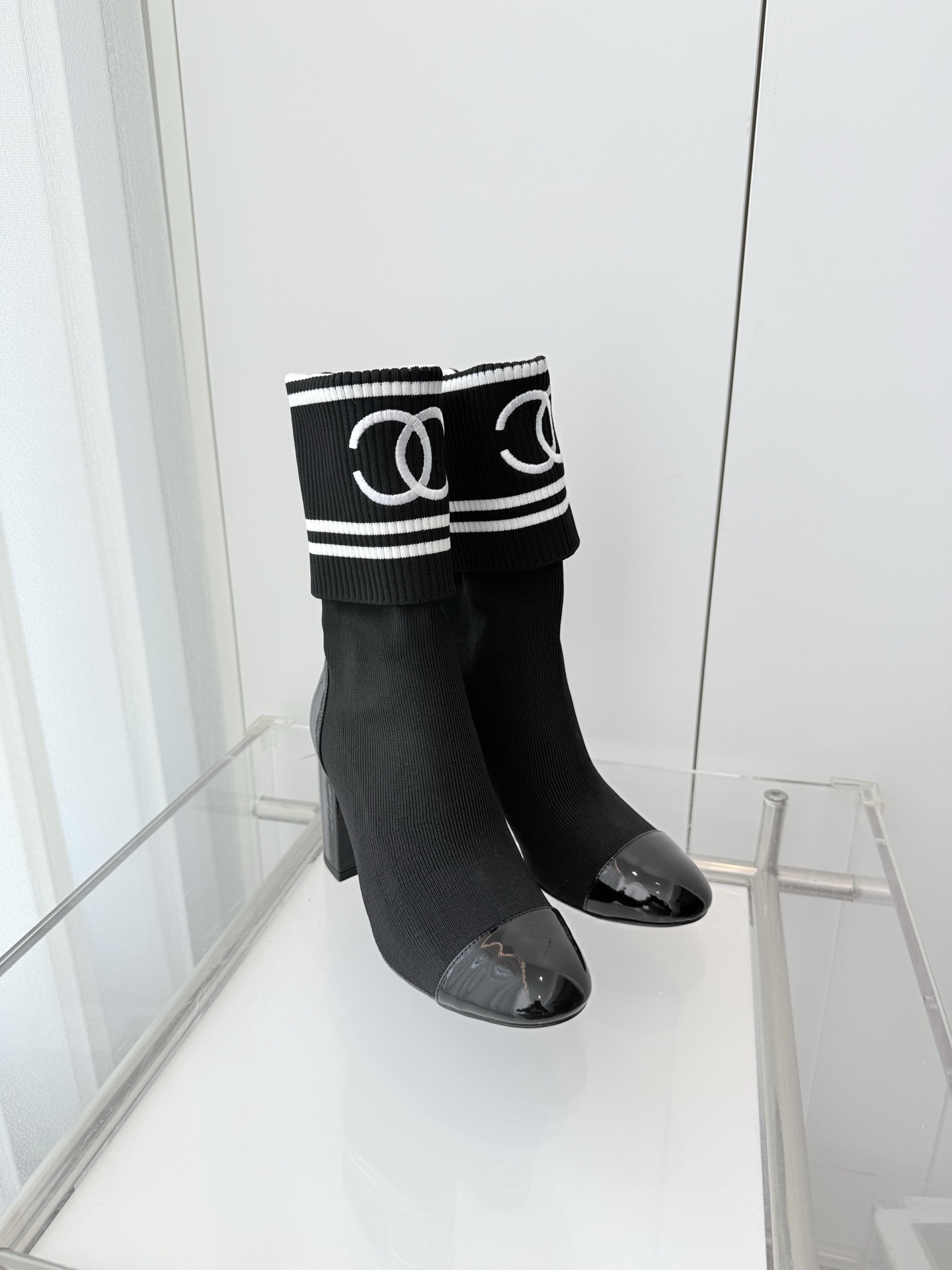 A pair of elegant black knit sock boots with patent leather cap toes and white logo fold-over cuffs displayed on a glass shelf. - Hình 4