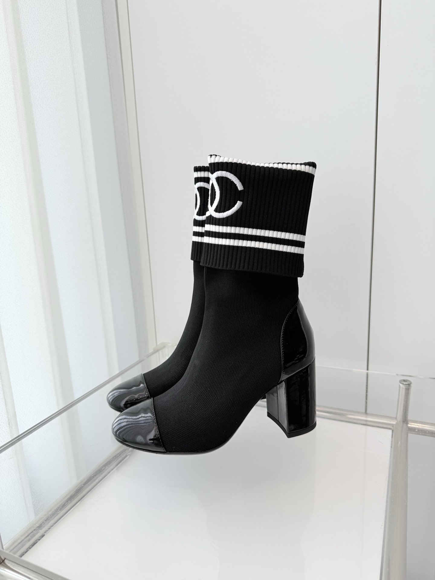 A pair of elegant black knit sock boots with patent leather cap toes and white logo fold-over cuffs displayed on a glass shelf. - Hình 3