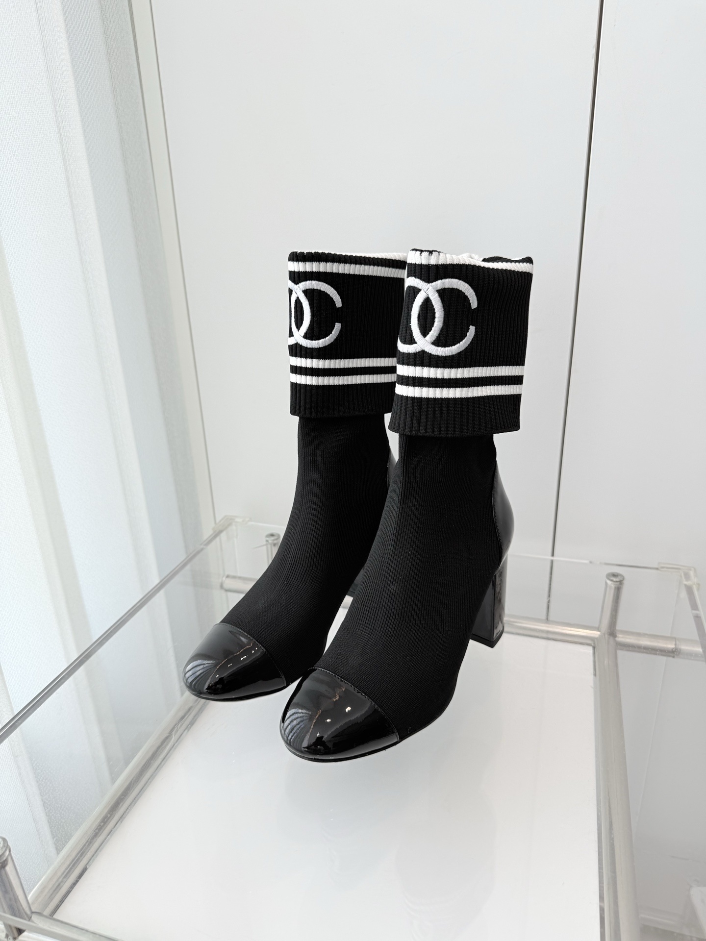 A pair of elegant black knit sock boots with patent leather cap toes and white logo fold-over cuffs displayed on a glass shelf. - Hình 2