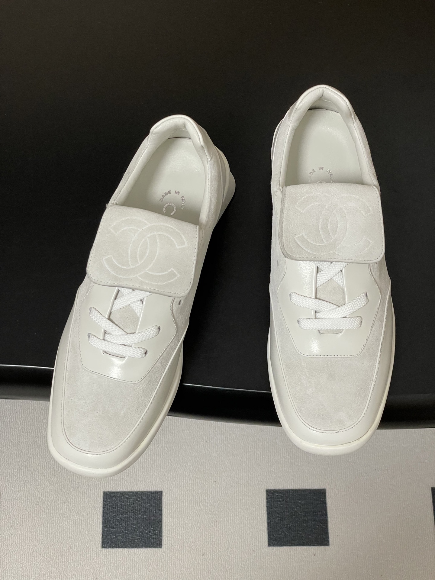 Pair of Chanel white leather and suede sneakers with embossed CC logo flap on a black surface - Hình 2