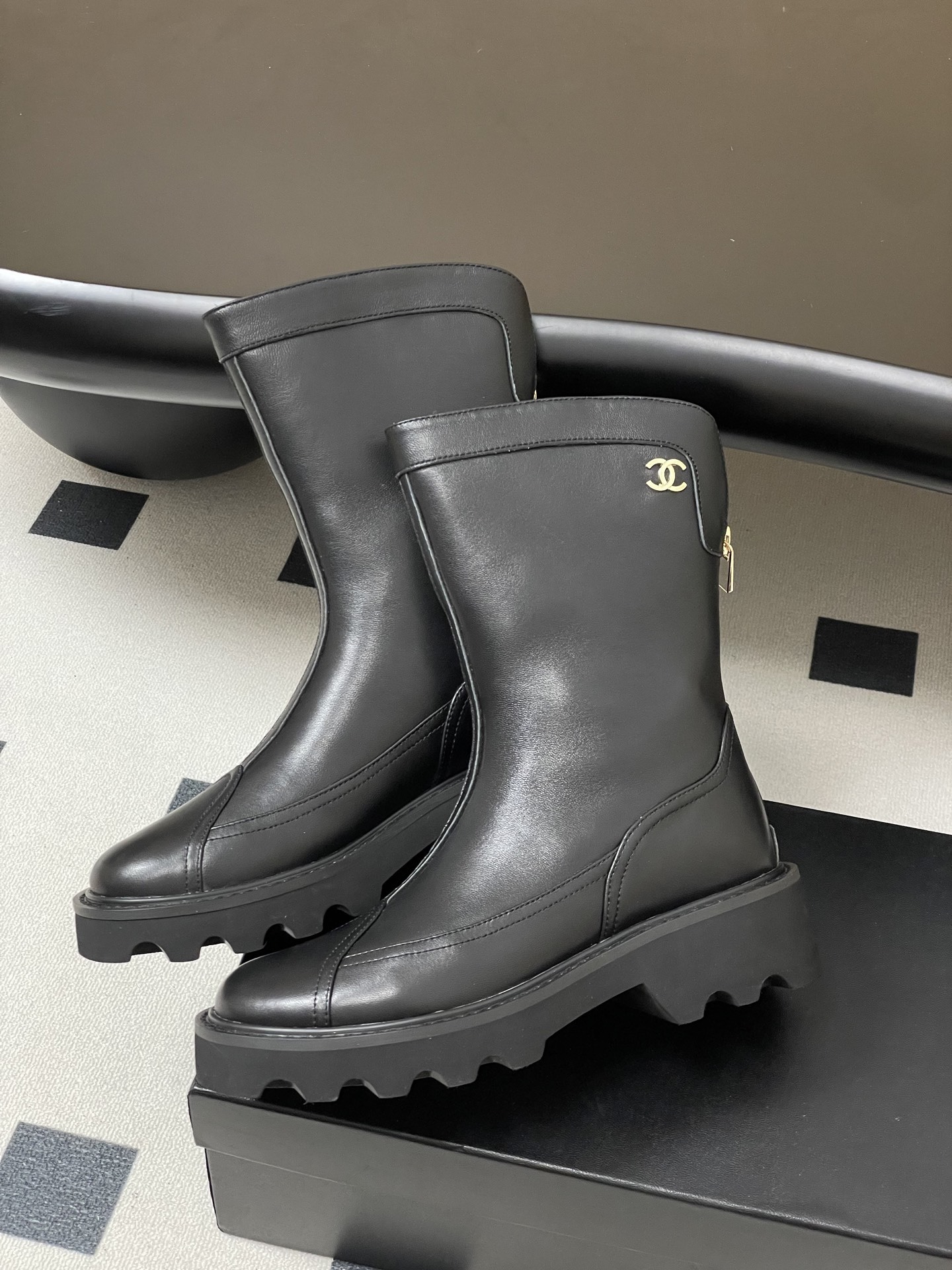 Pair of Chanel black leather mid-calf boots with chunky lug soles and gold CC logo hardware on a black surface. - Hình 4