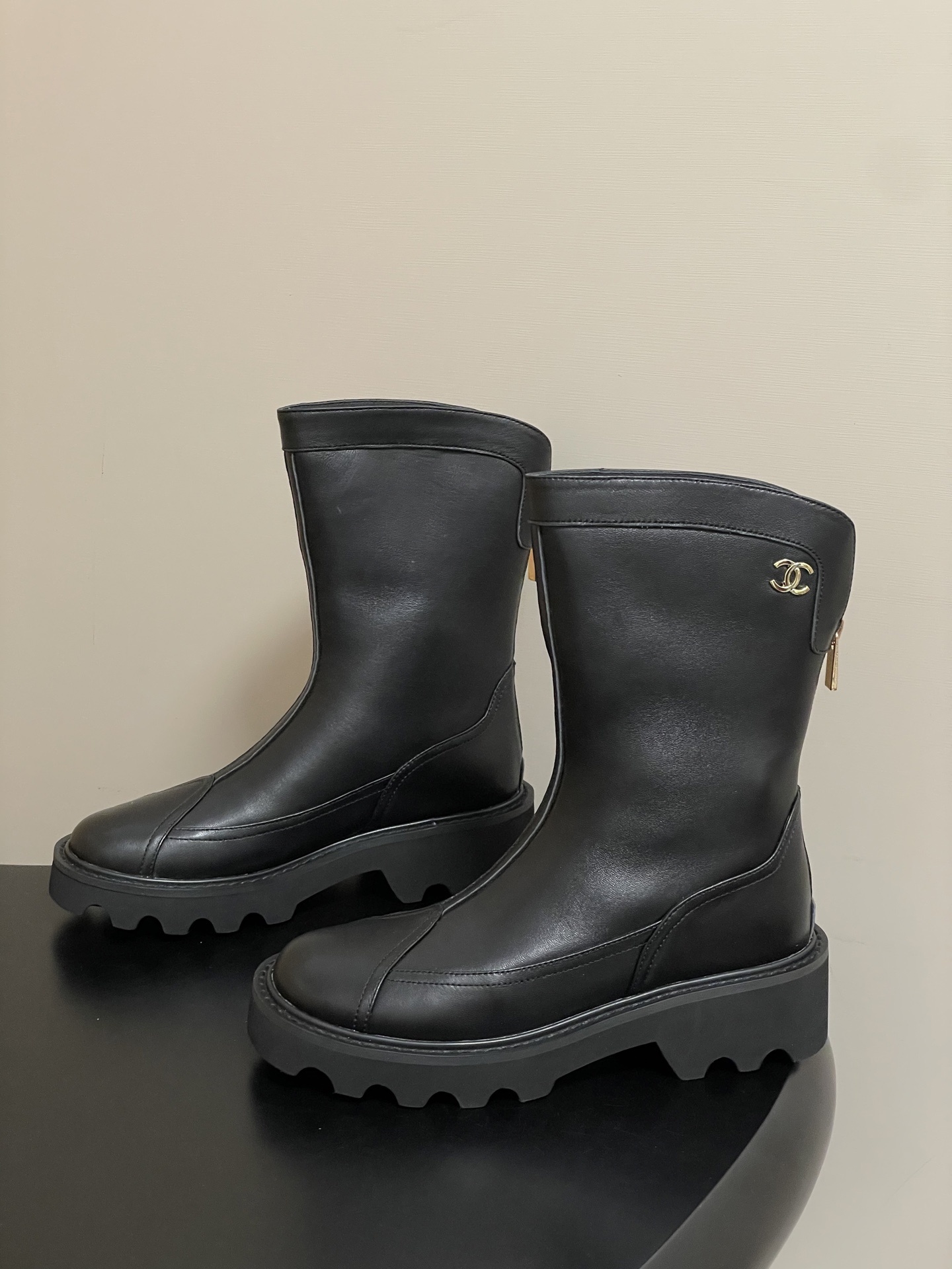 Pair of Chanel black leather mid-calf boots with chunky lug soles and gold CC logo hardware on a black surface. - Hình 3