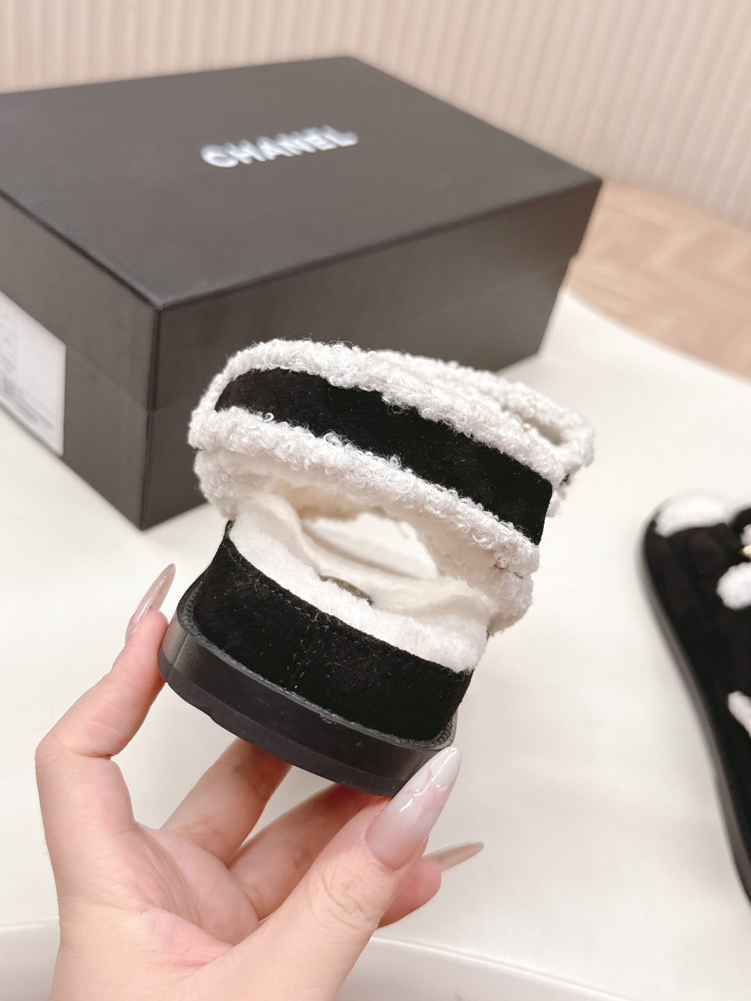 A pair of luxury black velvet sandals with white shearling trim and gold-tone logo buttons displayed on a white surface next to a black shoe box. - Hình 9