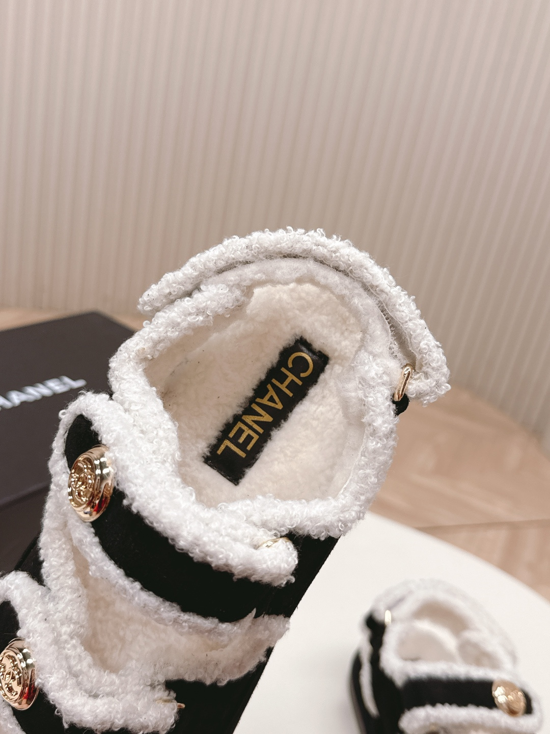 A pair of luxury black velvet sandals with white shearling trim and gold-tone logo buttons displayed on a white surface next to a black shoe box. - Hình 8