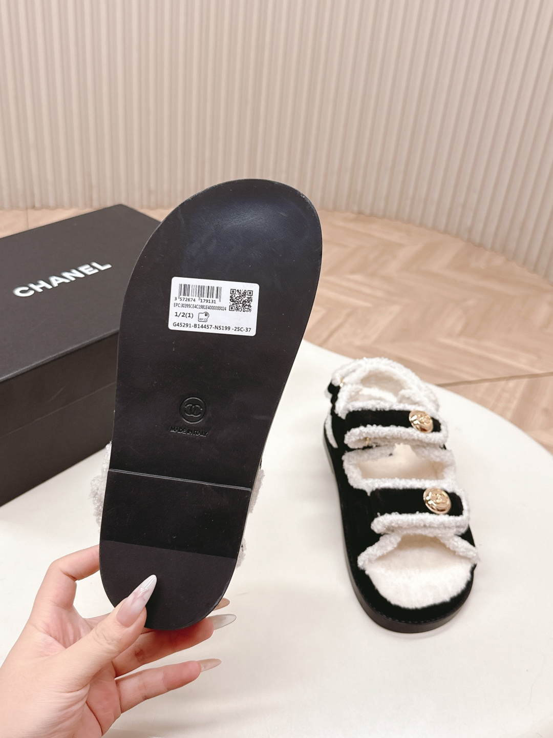 A pair of luxury black velvet sandals with white shearling trim and gold-tone logo buttons displayed on a white surface next to a black shoe box. - Hình 7