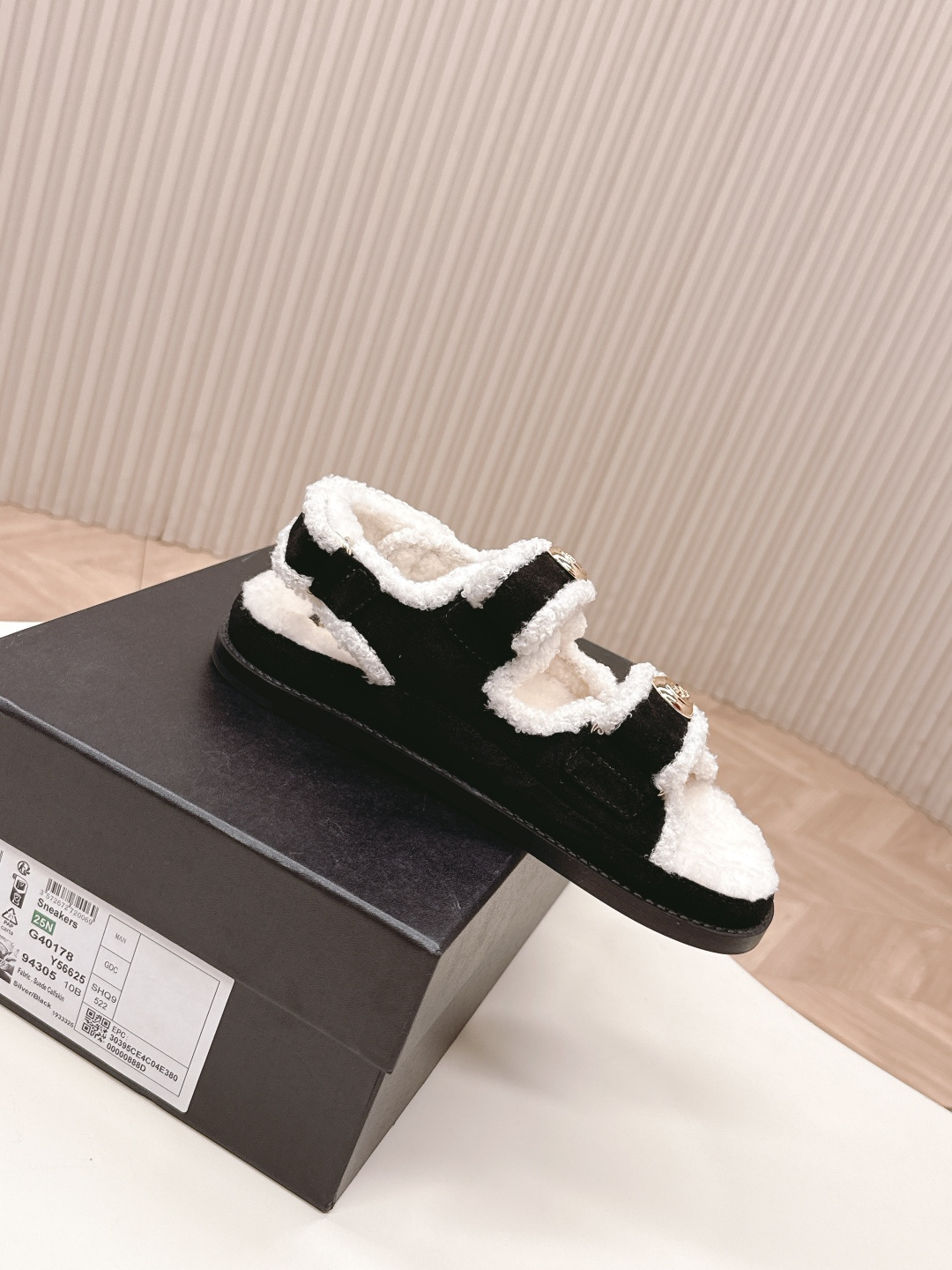A pair of luxury black velvet sandals with white shearling trim and gold-tone logo buttons displayed on a white surface next to a black shoe box. - Hình 5