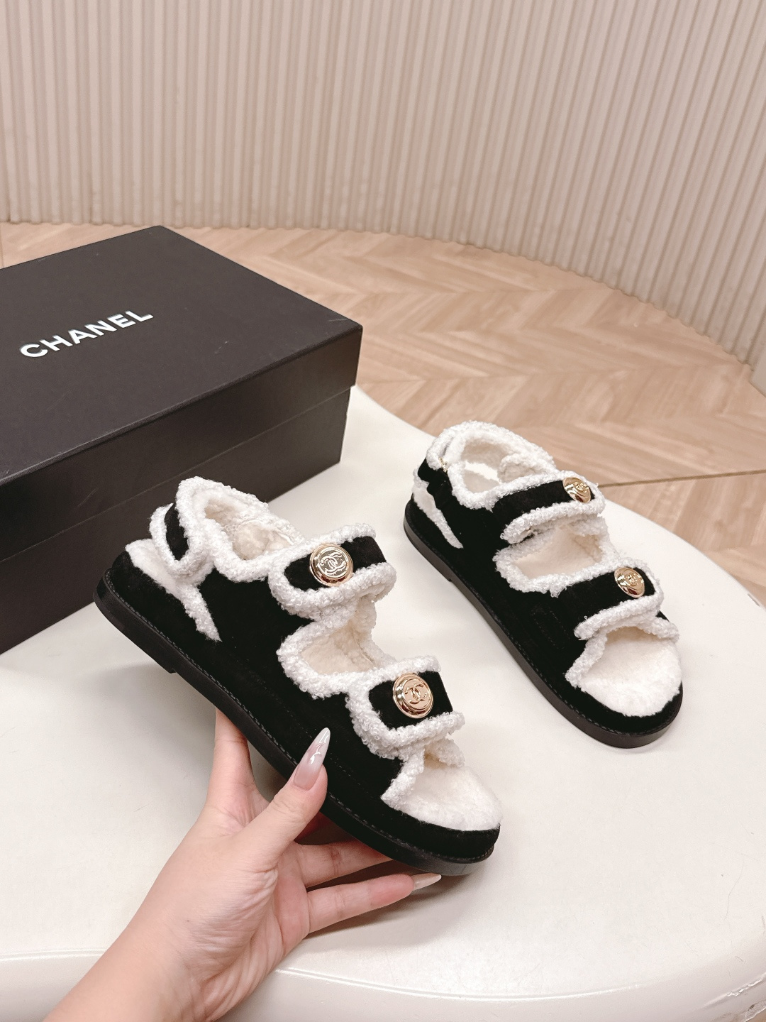 A pair of luxury black velvet sandals with white shearling trim and gold-tone logo buttons displayed on a white surface next to a black shoe box. - Hình 4