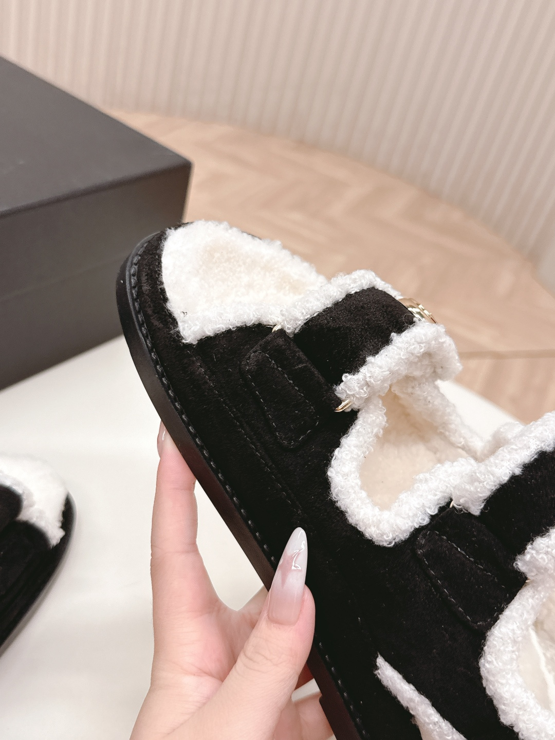 A pair of luxury black velvet sandals with white shearling trim and gold-tone logo buttons displayed on a white surface next to a black shoe box. - Hình 3