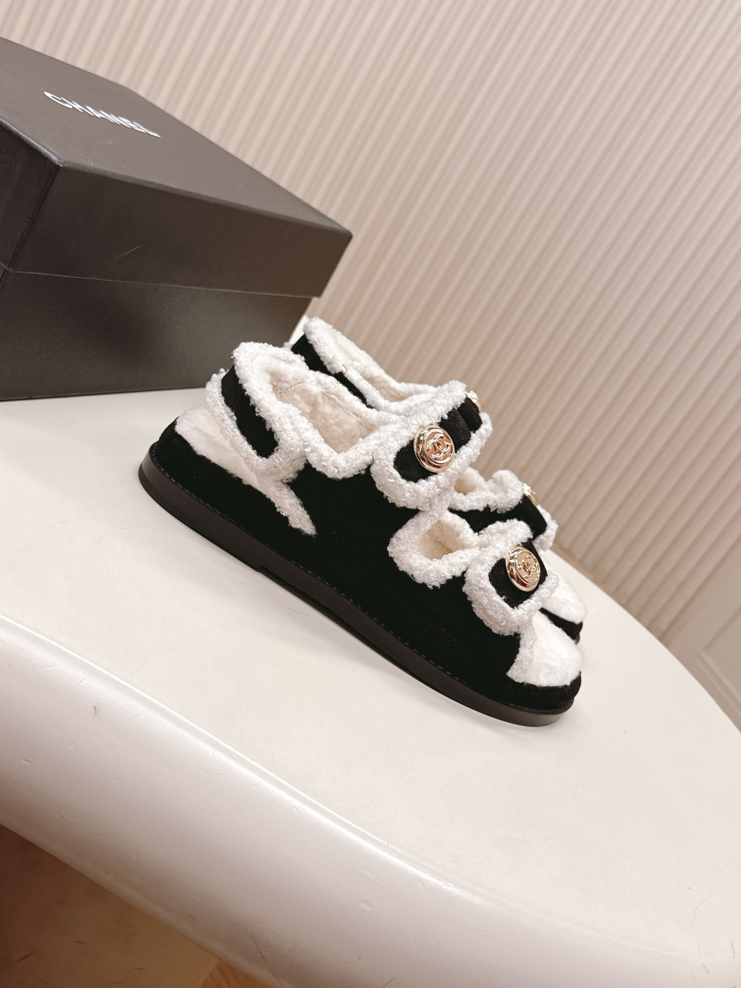 A pair of luxury black velvet sandals with white shearling trim and gold-tone logo buttons displayed on a white surface next to a black shoe box. - Hình 2
