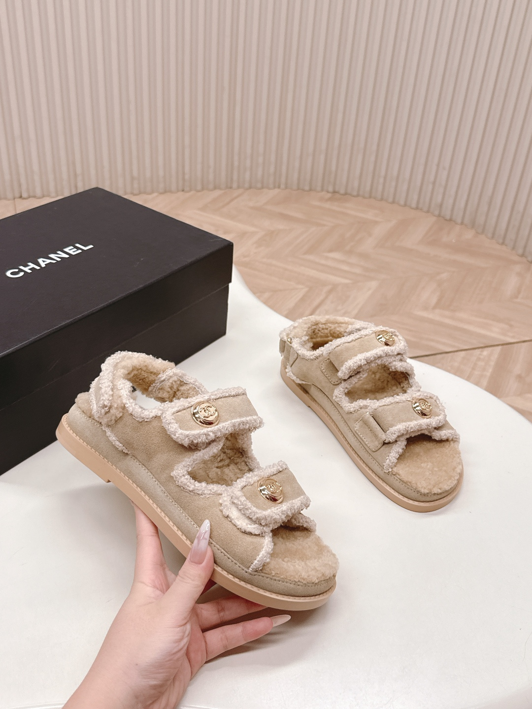 A pair of Chanel beige shearling dad sandals with gold CC logo buttons next to a black Chanel box. - Hình 8
