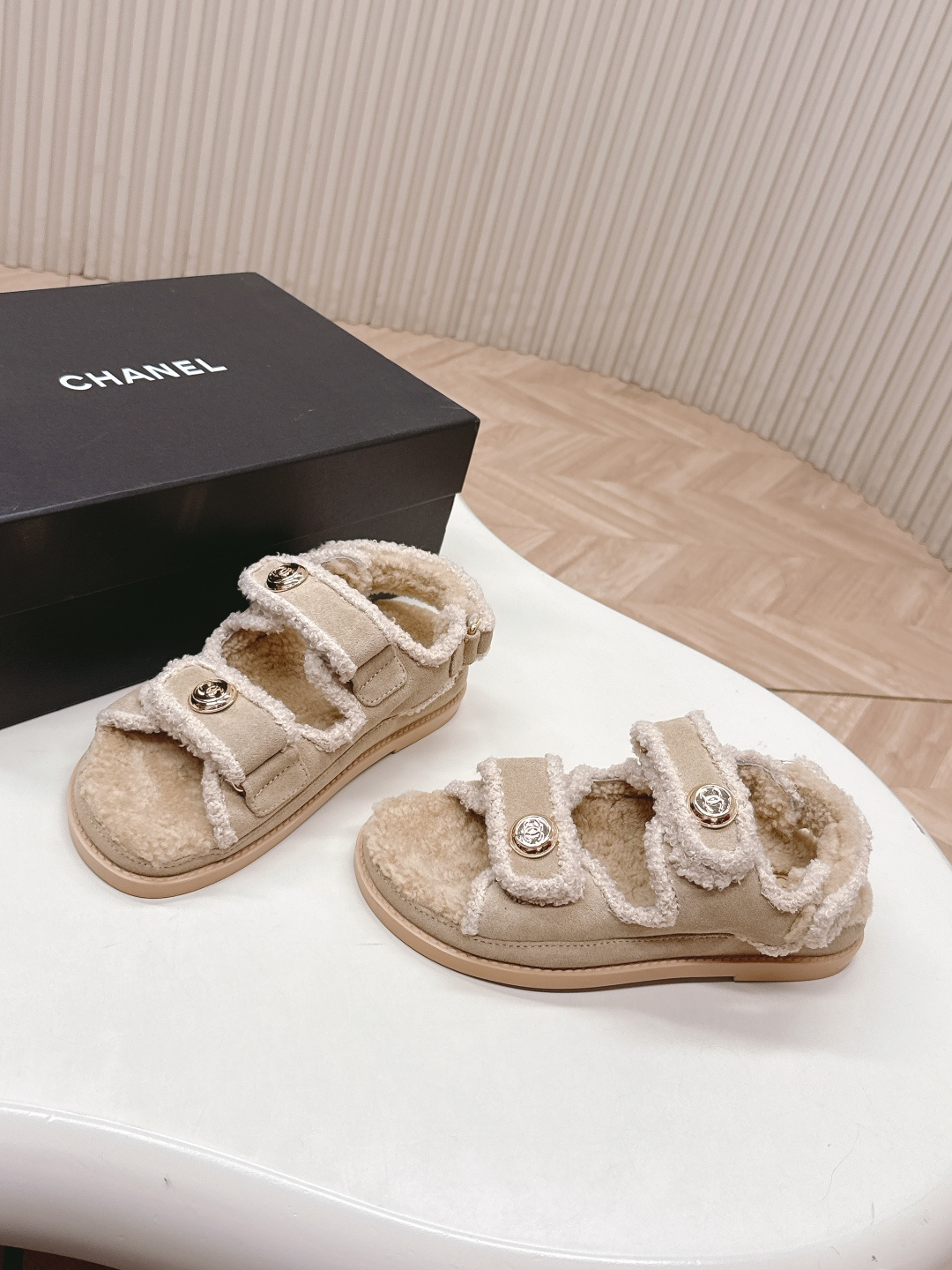A pair of Chanel beige shearling dad sandals with gold CC logo buttons next to a black Chanel box. - Hình 3