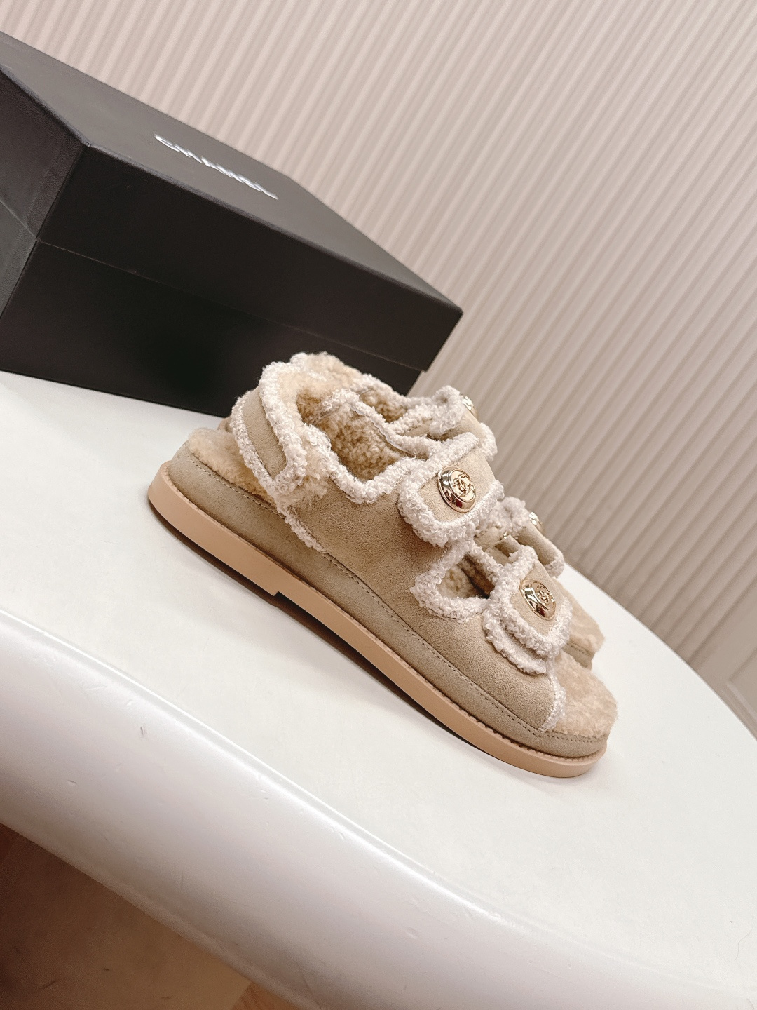 A pair of Chanel beige shearling dad sandals with gold CC logo buttons next to a black Chanel box. - Hình 2