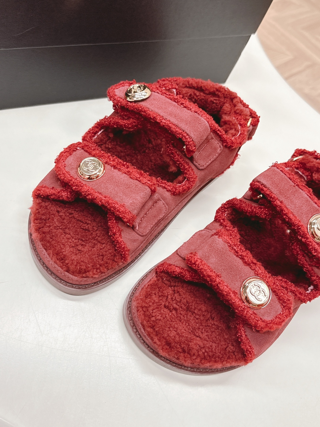Luxury burgundy shearling sandals with gold logo buttons displayed on a white surface next to a brand box. - Hình 5