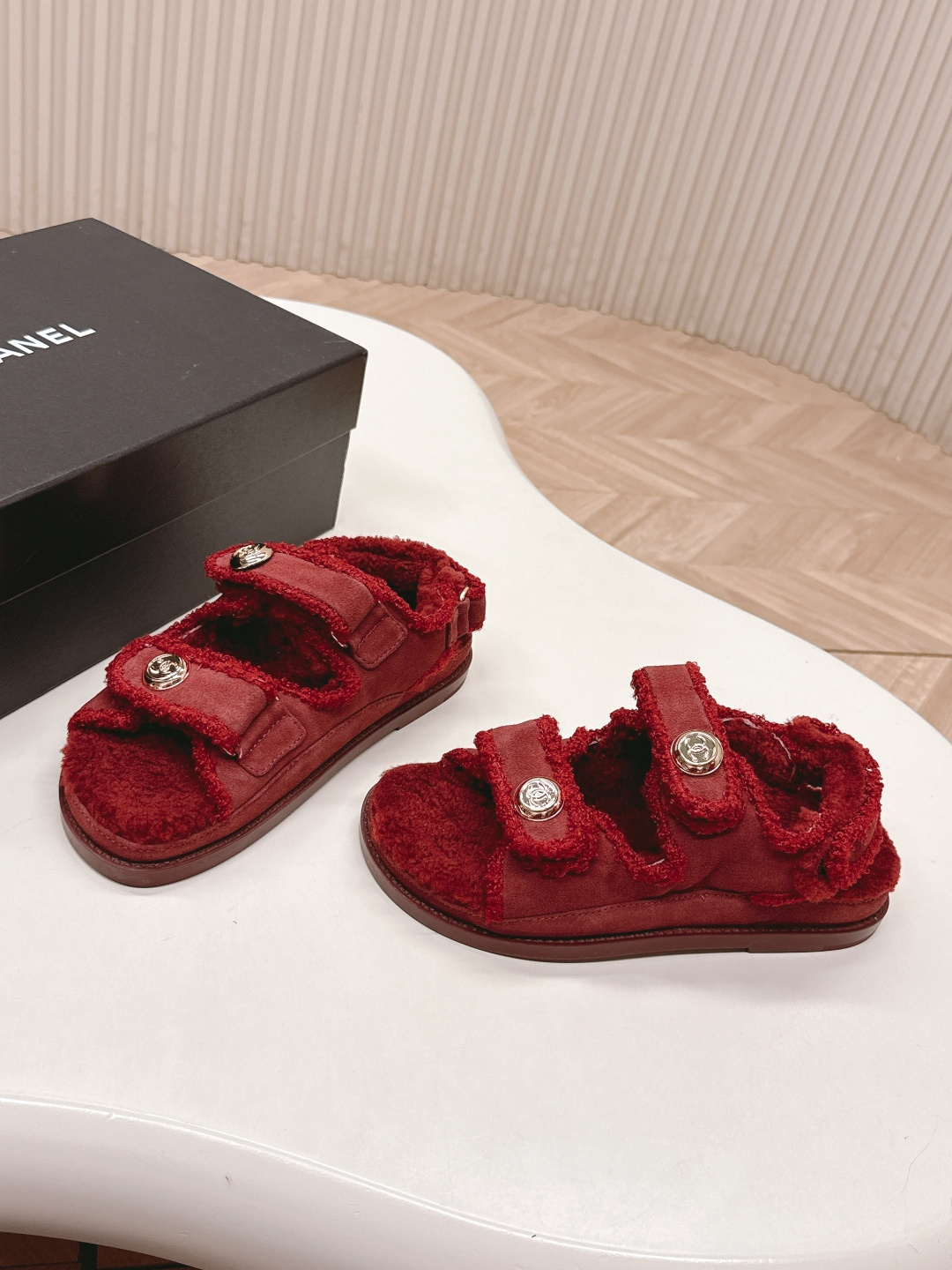 Luxury burgundy shearling sandals with gold logo buttons displayed on a white surface next to a brand box. - Hình 3