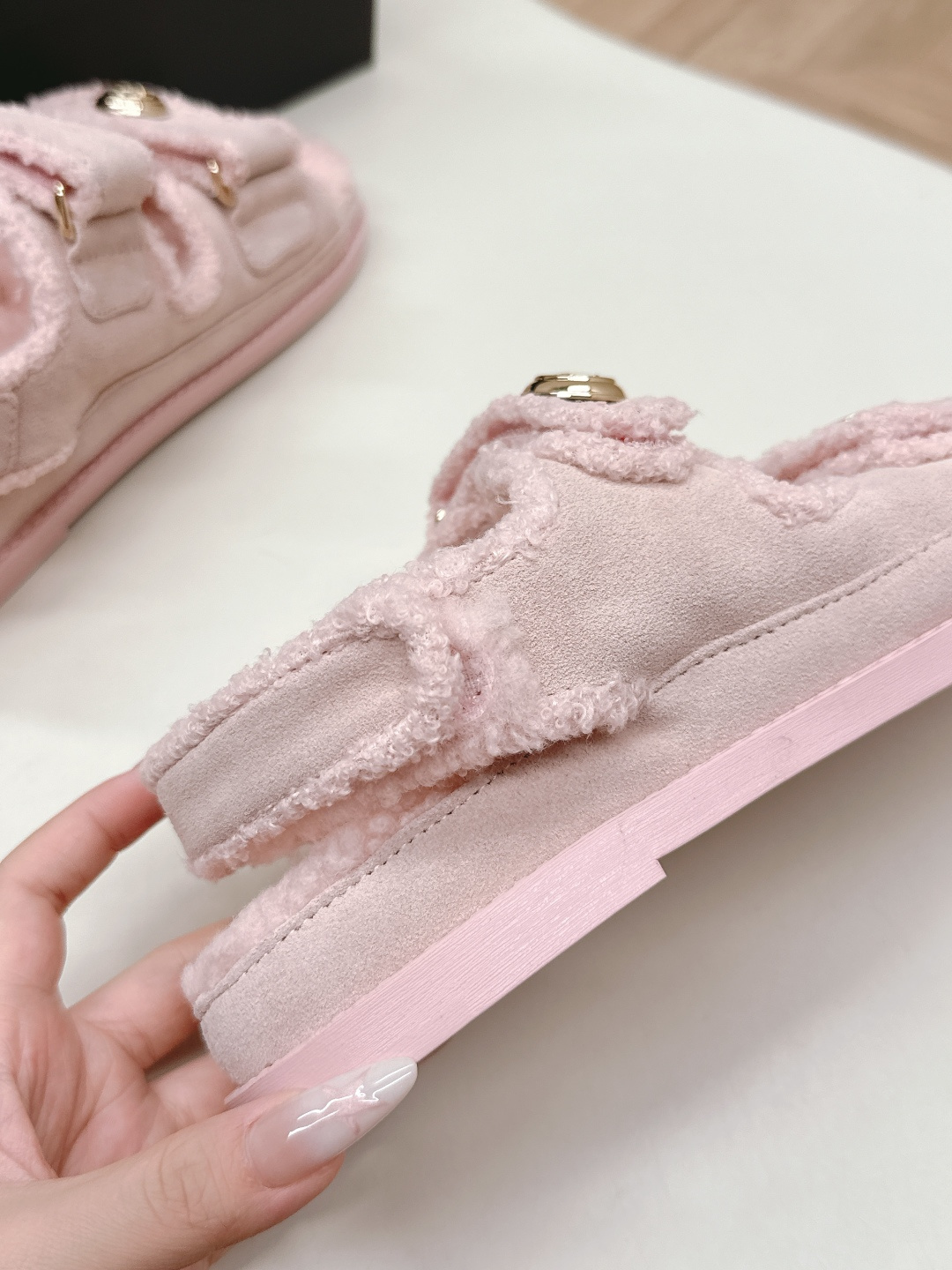 A pair of light pink fuzzy shearling sandals with gold double C logo buttons on a white surface next to a black designer box. - Hình 9