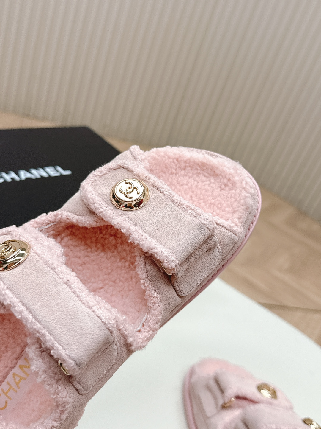 A pair of light pink fuzzy shearling sandals with gold double C logo buttons on a white surface next to a black designer box. - Hình 8