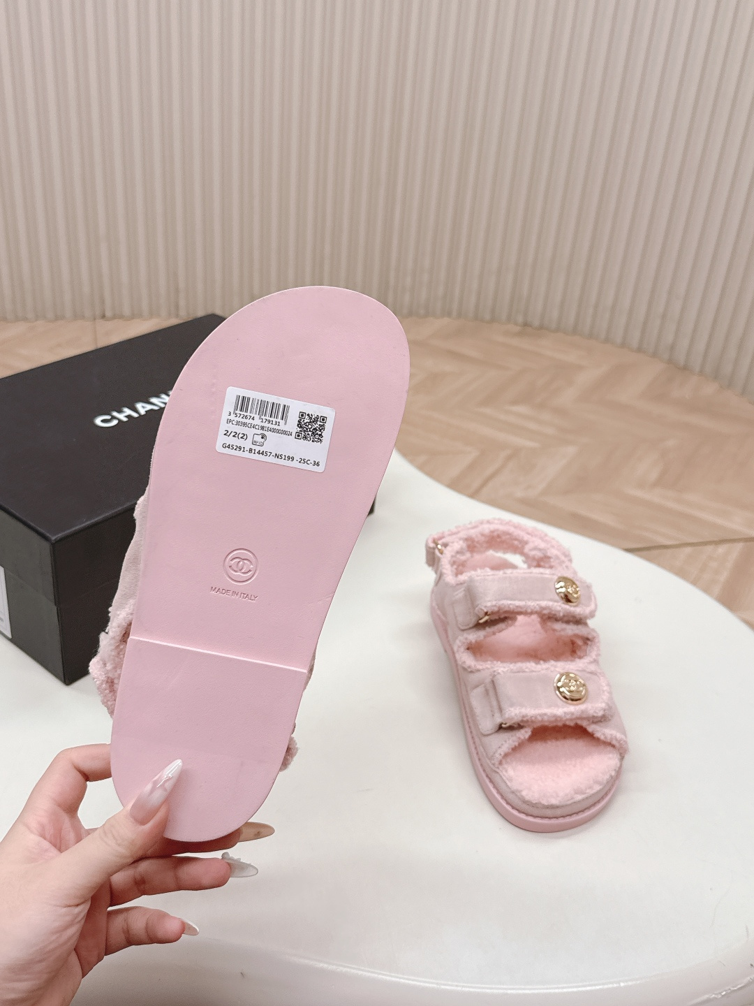 A pair of light pink fuzzy shearling sandals with gold double C logo buttons on a white surface next to a black designer box. - Hình 7