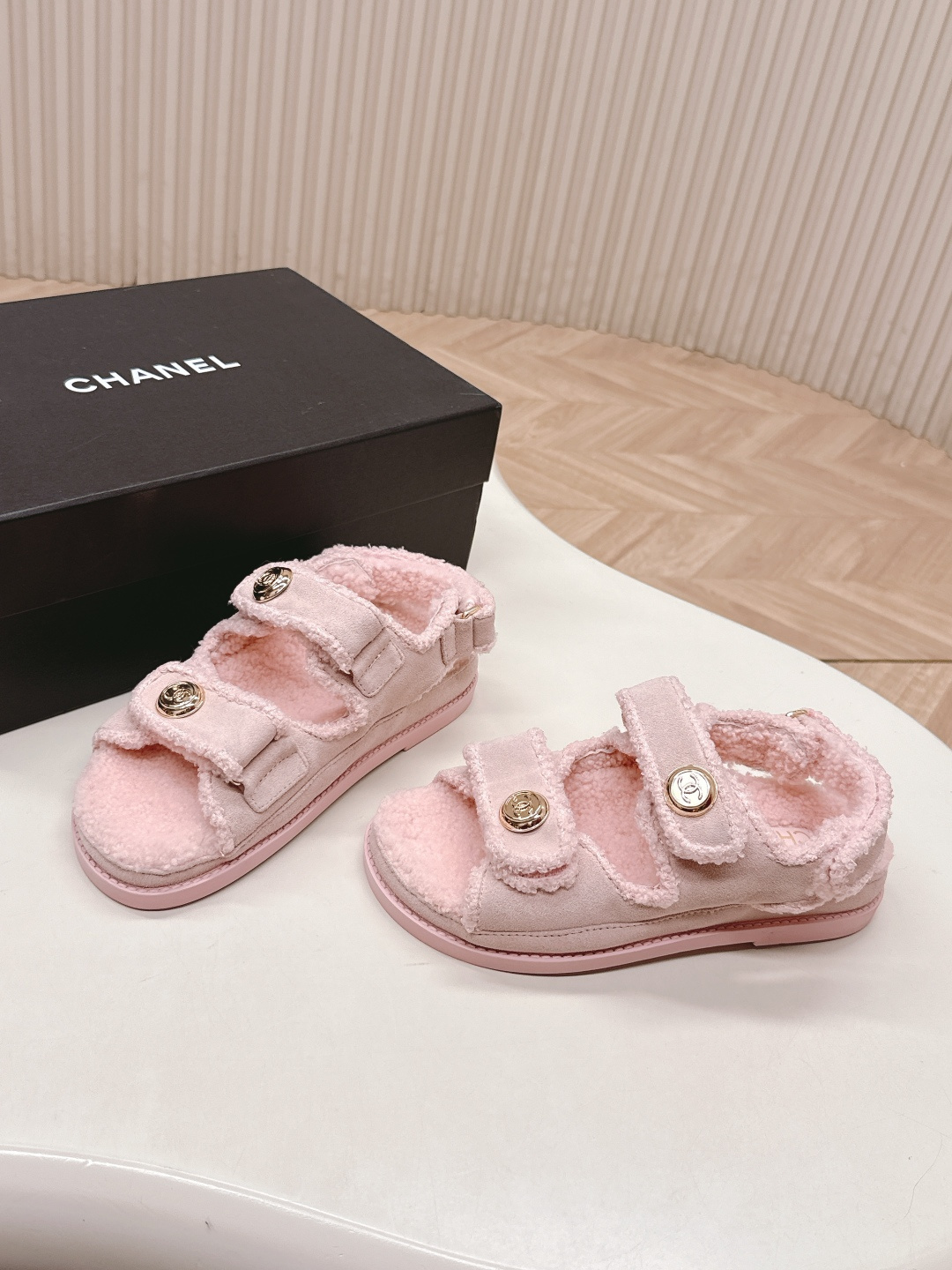 A pair of light pink fuzzy shearling sandals with gold double C logo buttons on a white surface next to a black designer box. - Hình 6