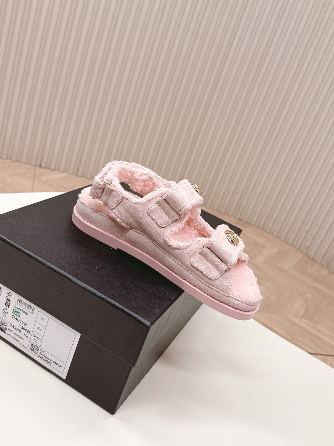 A pair of light pink fuzzy shearling sandals with gold double C logo buttons on a white surface next to a black designer box. - Hình 5
