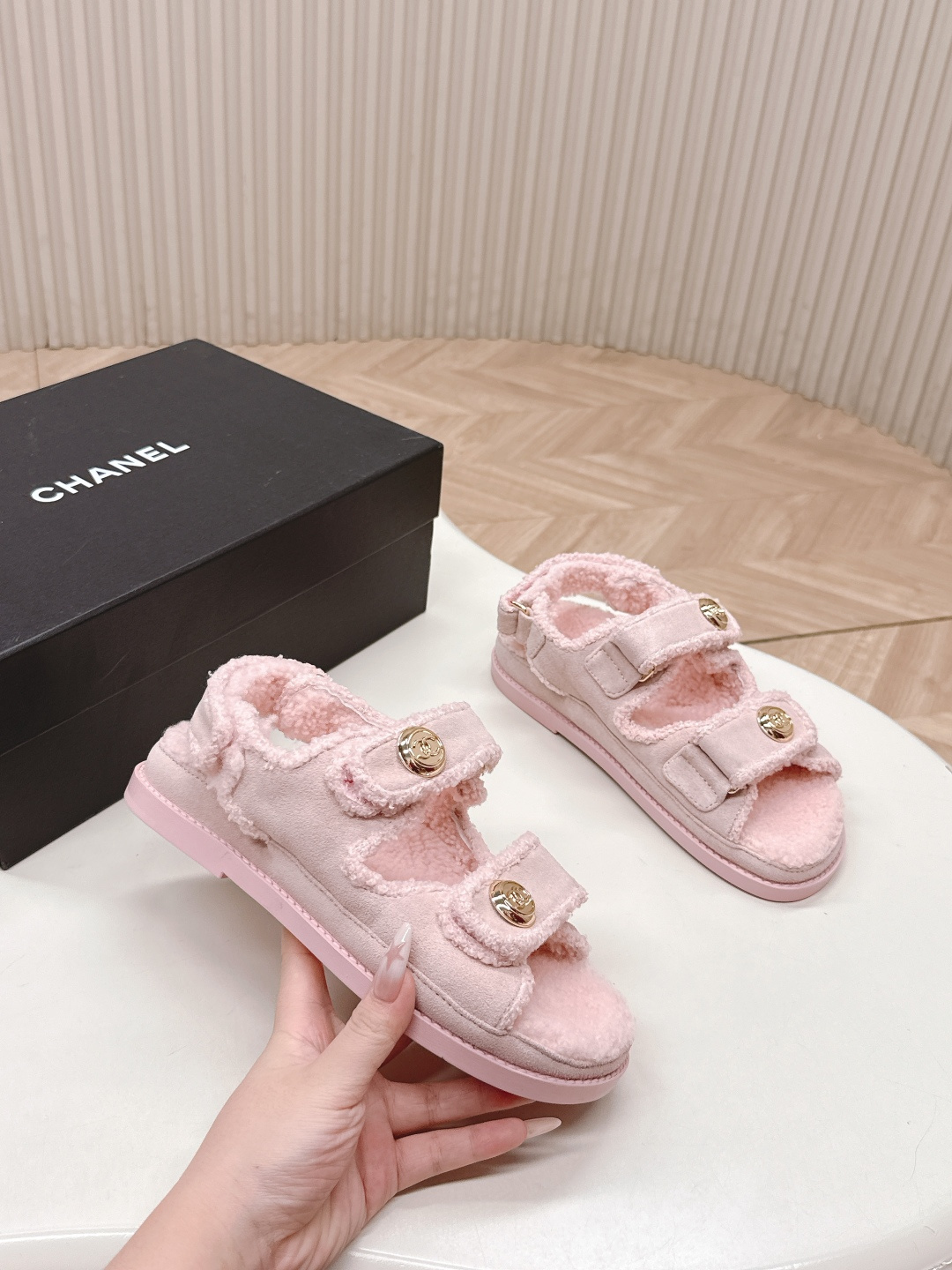 A pair of light pink fuzzy shearling sandals with gold double C logo buttons on a white surface next to a black designer box. - Hình 4
