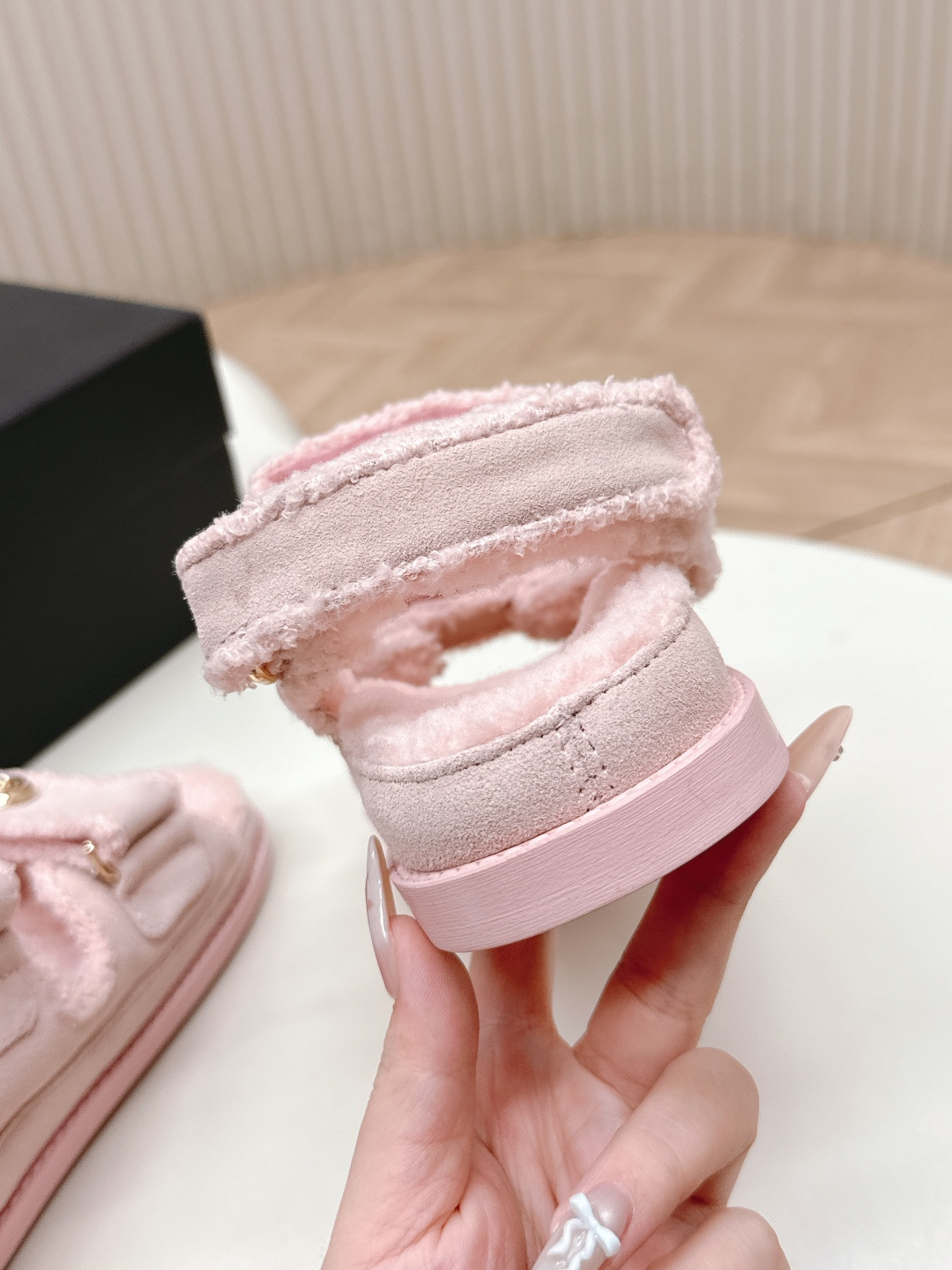 A pair of light pink fuzzy shearling sandals with gold double C logo buttons on a white surface next to a black designer box. - Hình 3