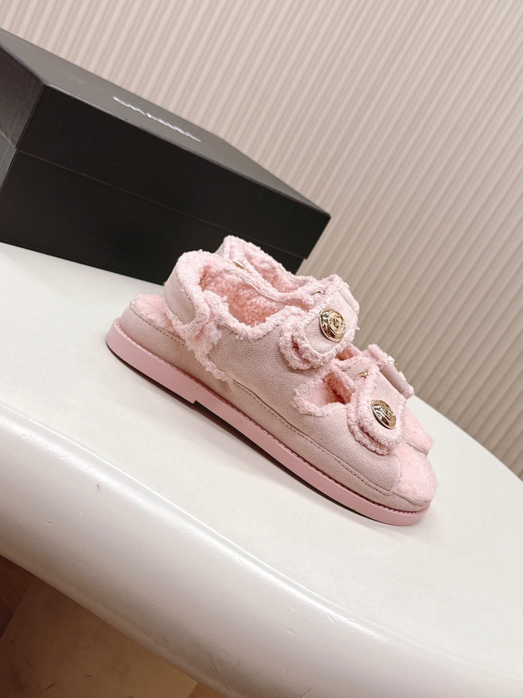 A pair of light pink fuzzy shearling sandals with gold double C logo buttons on a white surface next to a black designer box. - Hình 2