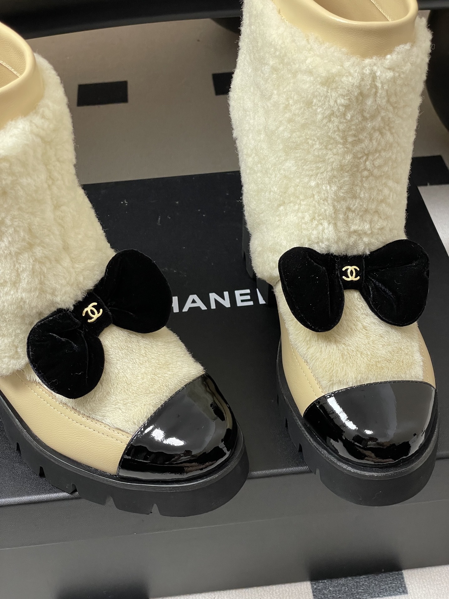 Pair of cream Chanel shearling ankle boots with black patent toe caps and velvet bows on a black table - Hình 9