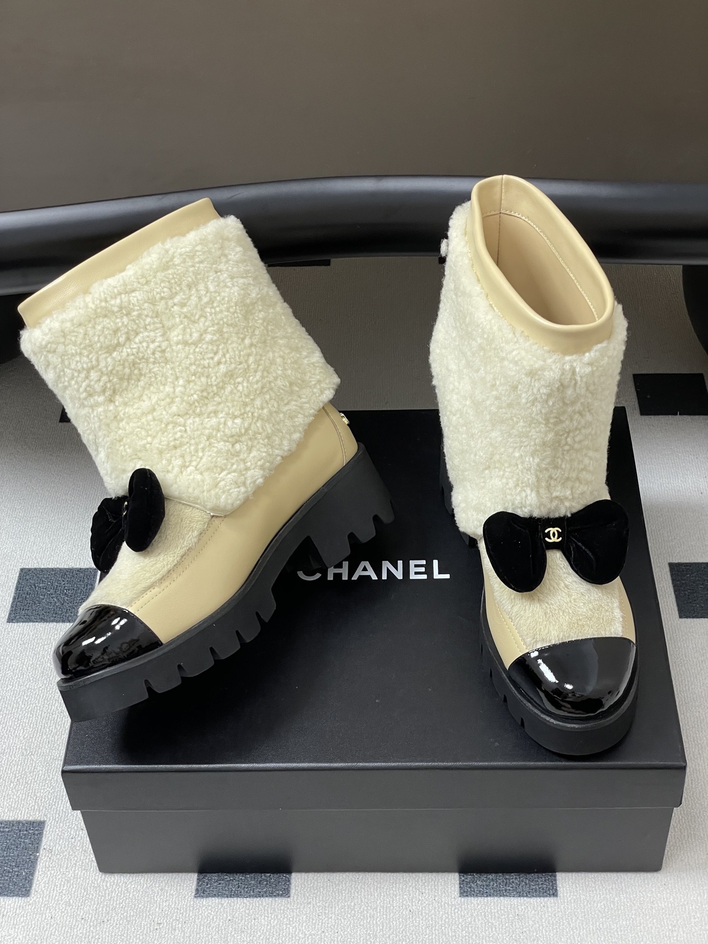 Pair of cream Chanel shearling ankle boots with black patent toe caps and velvet bows on a black table - Hình 5