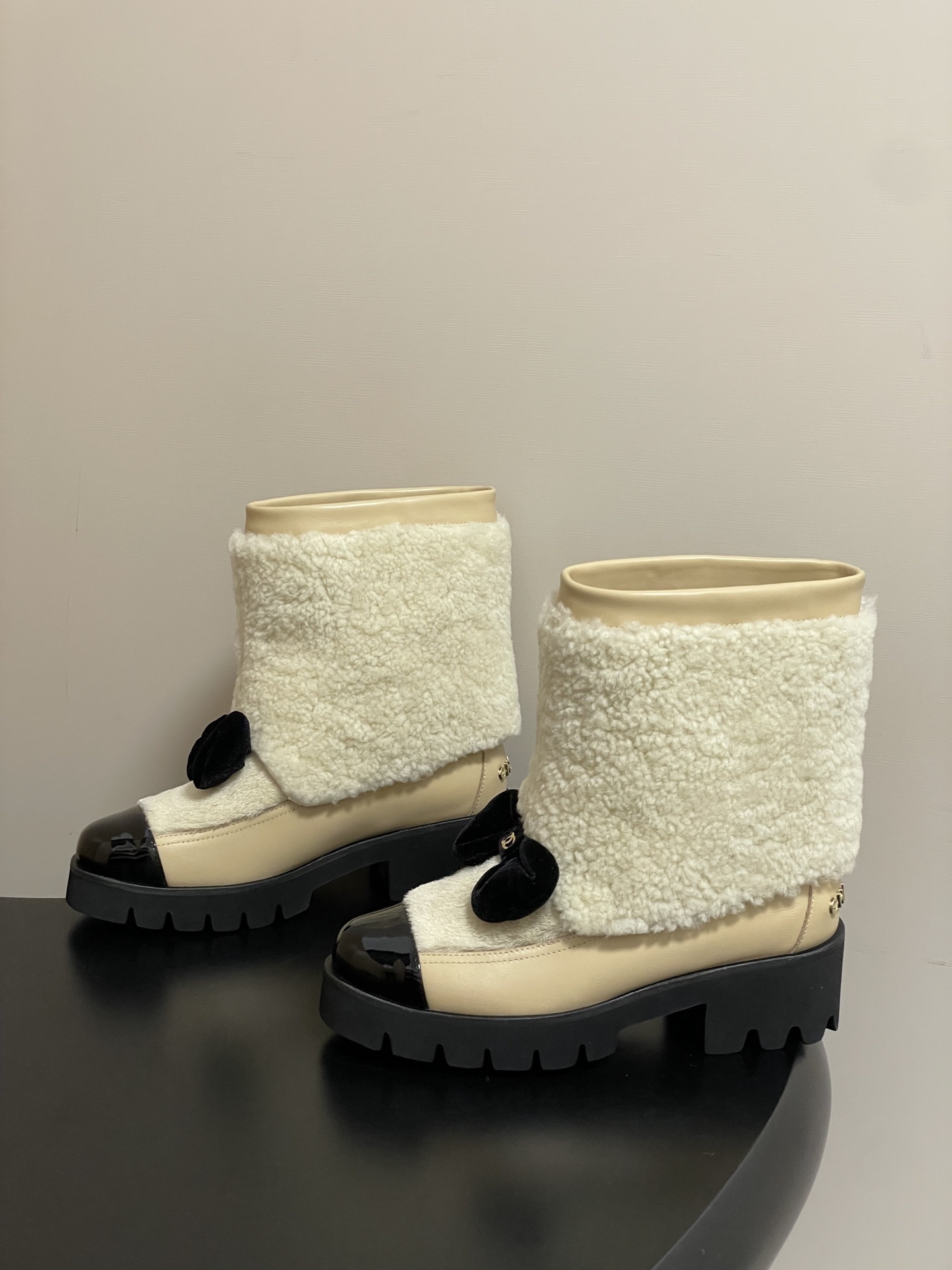 Pair of cream Chanel shearling ankle boots with black patent toe caps and velvet bows on a black table - Hình 3