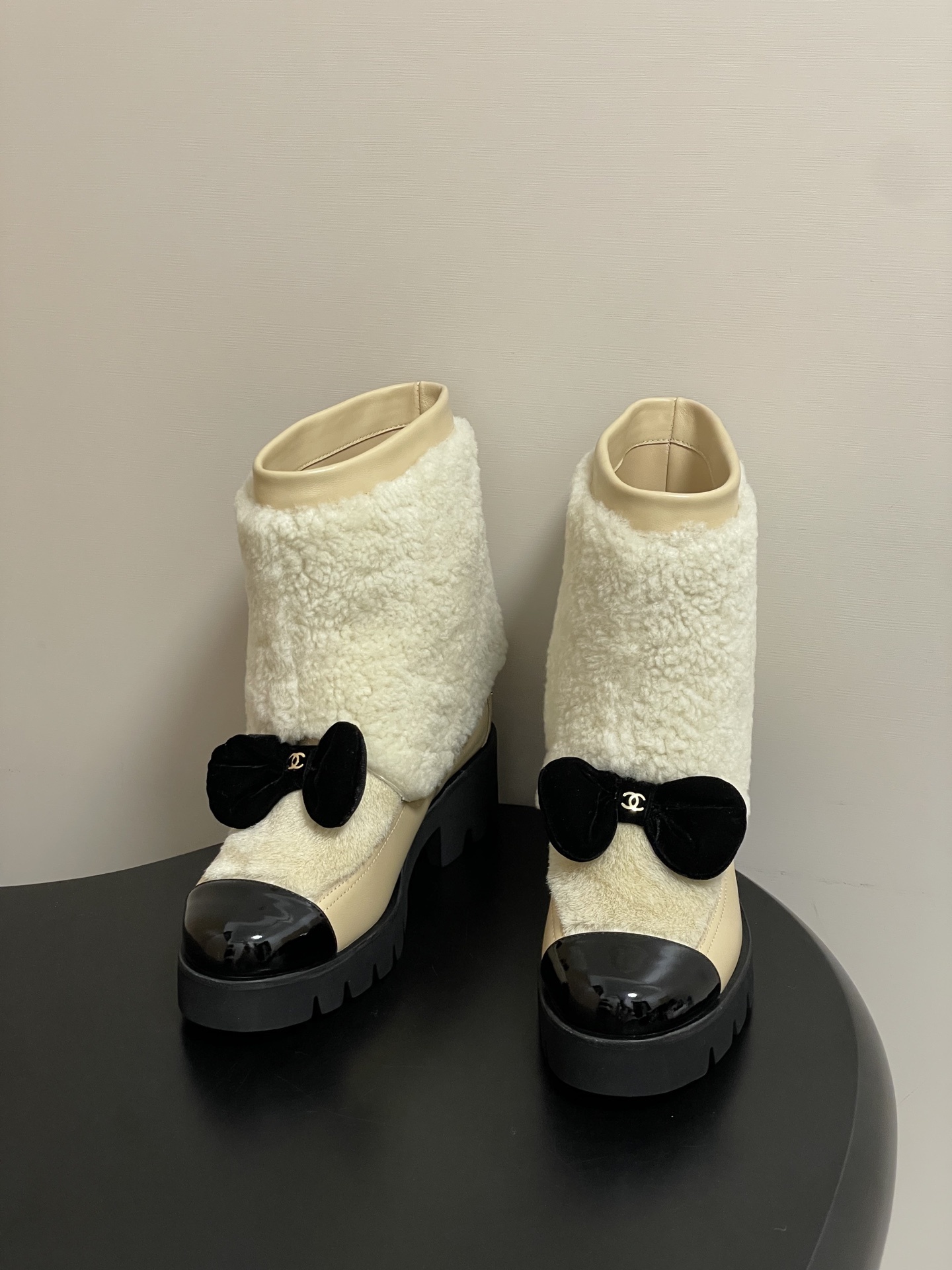 Pair of cream Chanel shearling ankle boots with black patent toe caps and velvet bows on a black table - Hình 2