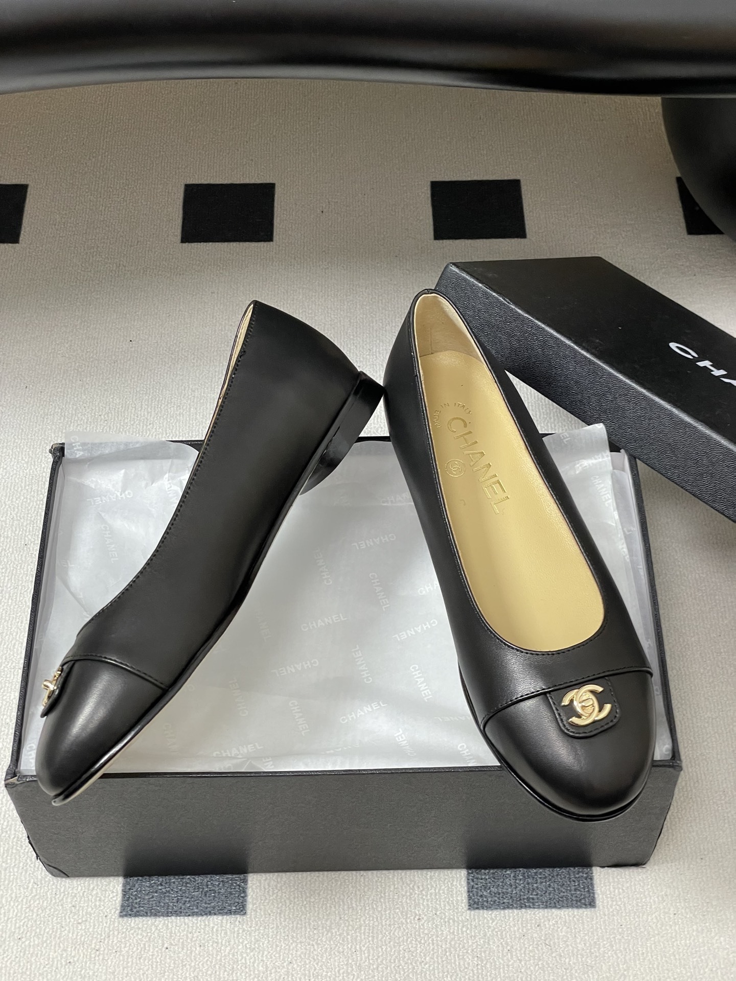 A pair of luxury Chanel black lambskin ballerina flats featuring a gold CC logo on the toe cap, displayed on a black surface. - Hình 6