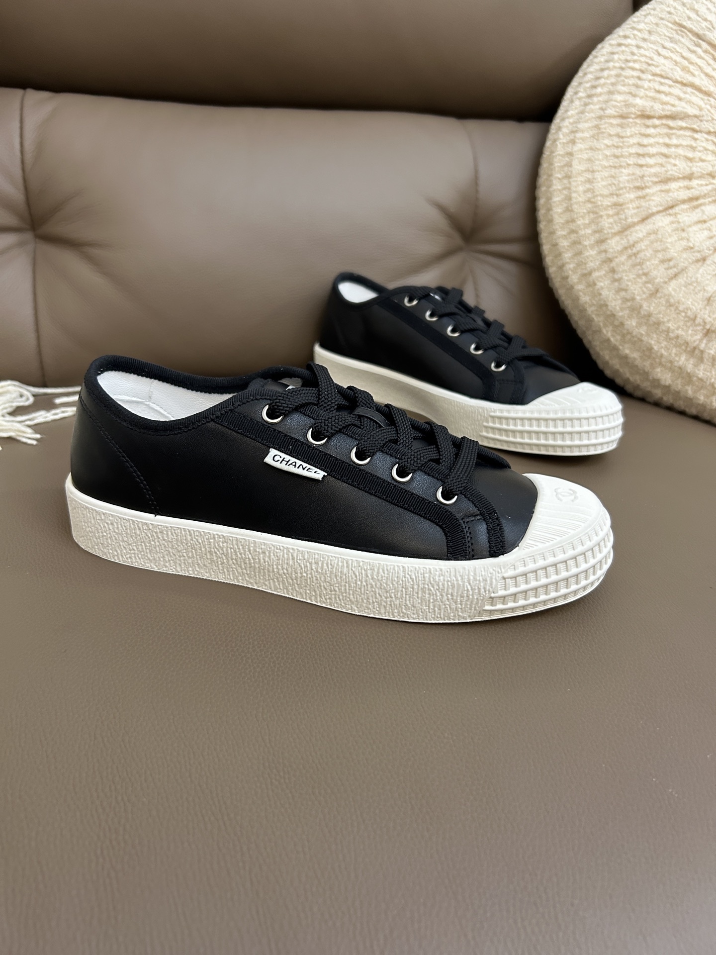 Pair of Chanel luxury black leather low-top sneakers with white rubber soles and toe caps on a brown leather background. - Hình 3