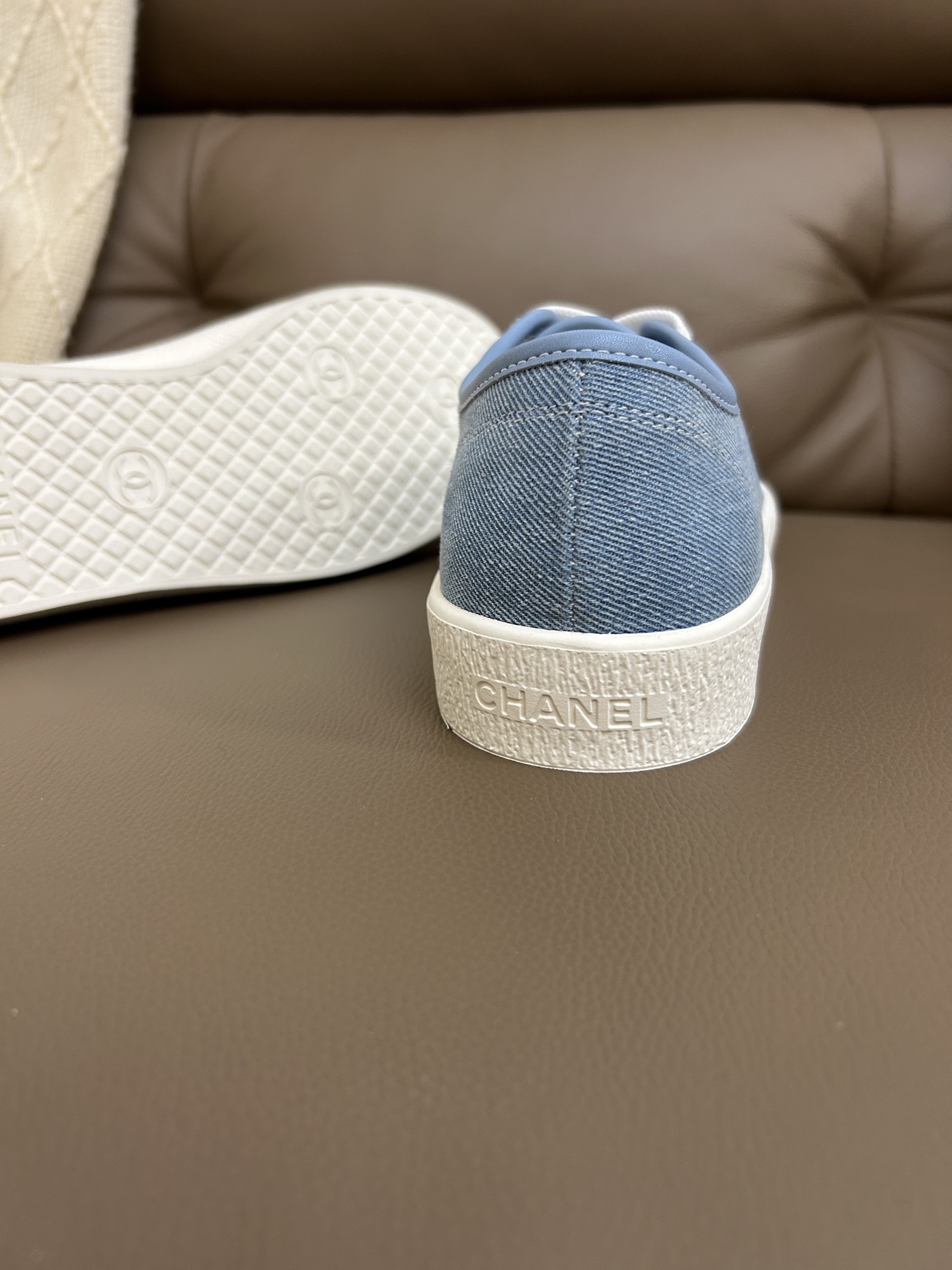 A pair of luxury light blue Chanel denim low-top sneakers with white rubber soles and white laces on a brown leather surface. - Hình 9