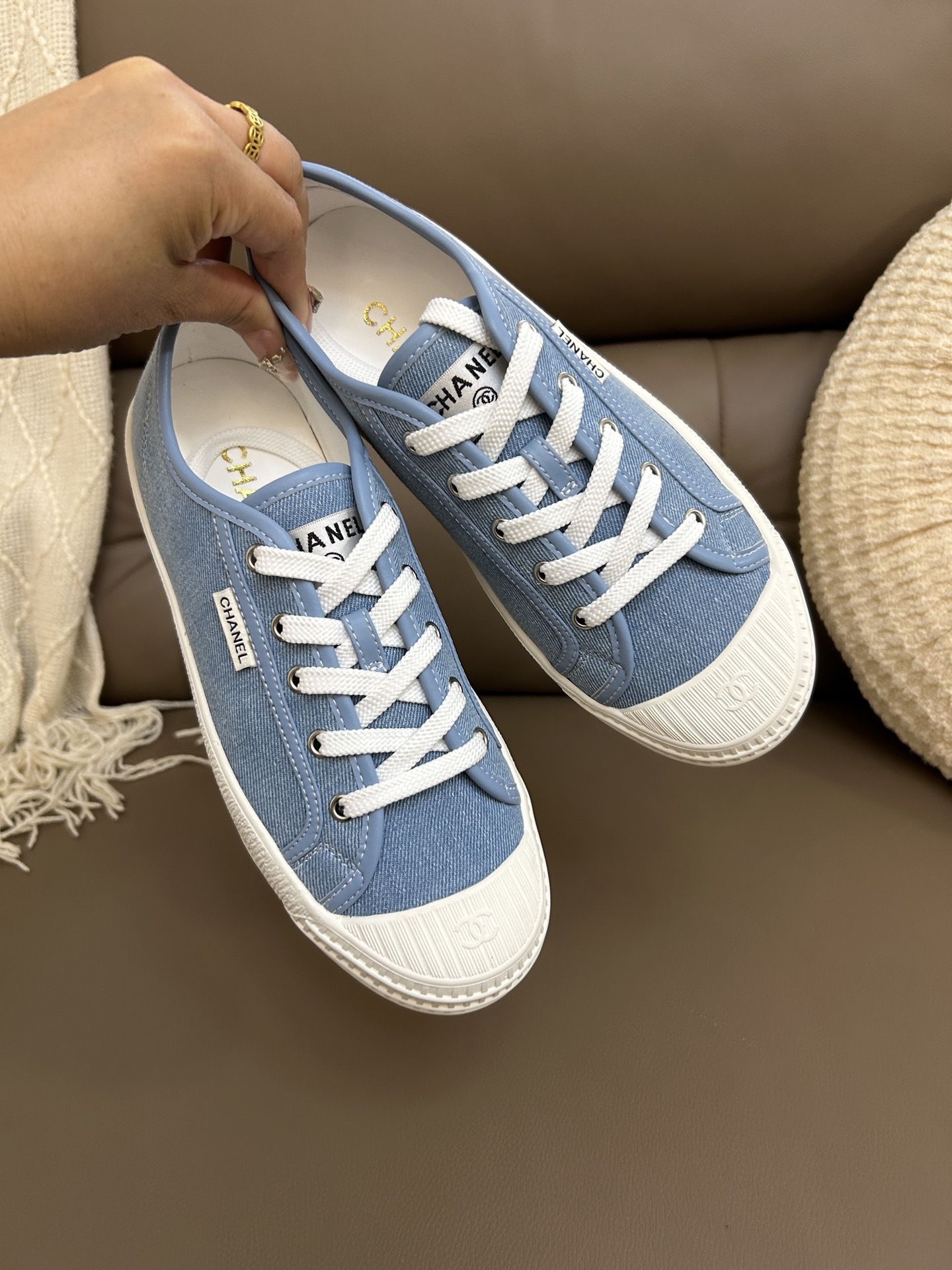 A pair of luxury light blue Chanel denim low-top sneakers with white rubber soles and white laces on a brown leather surface. - Hình 2