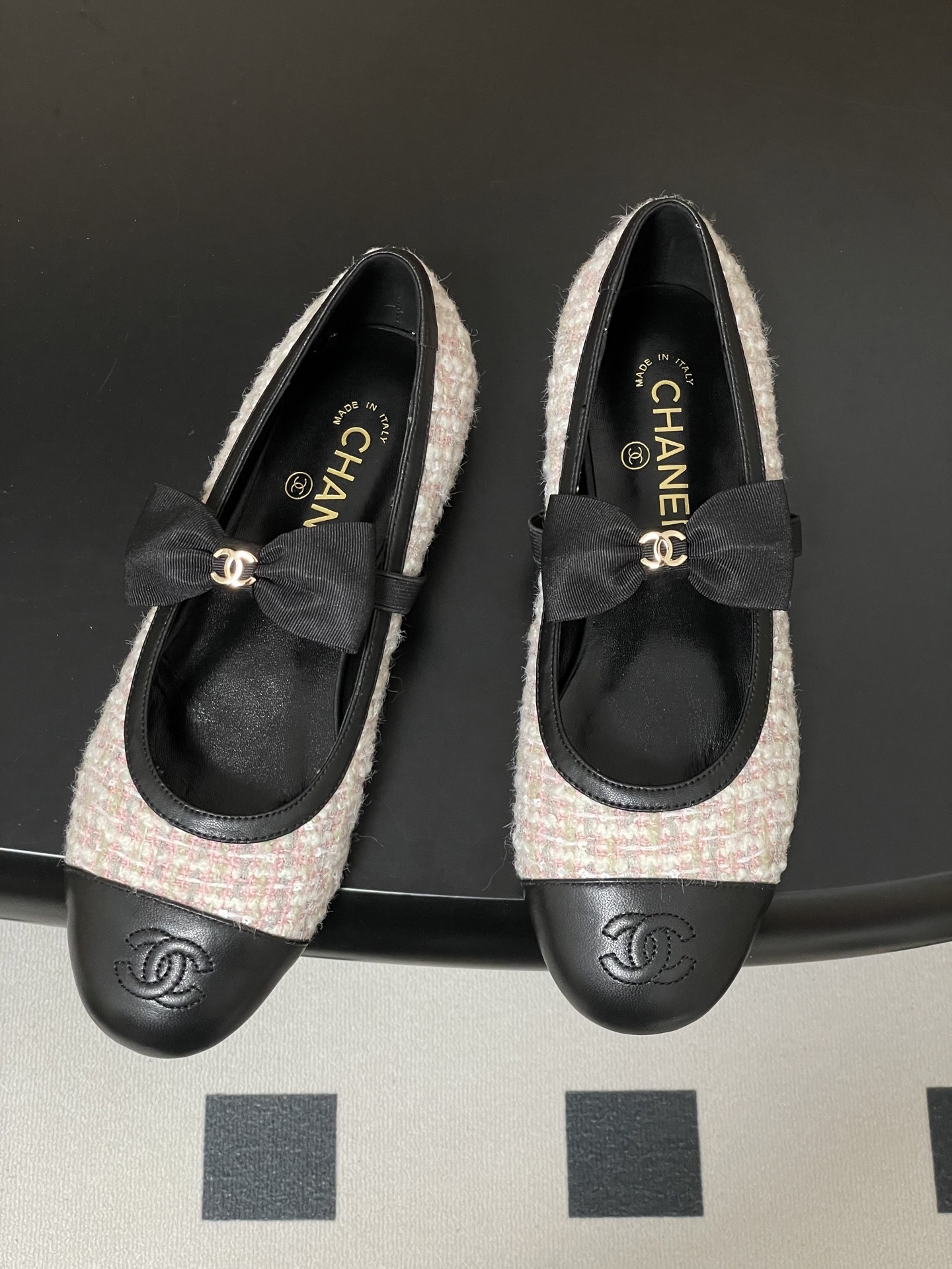 Pair of Chanel pink and white tweed ballet flats with black leather cap toes and CC logo bows on a black surface. - Hình 2