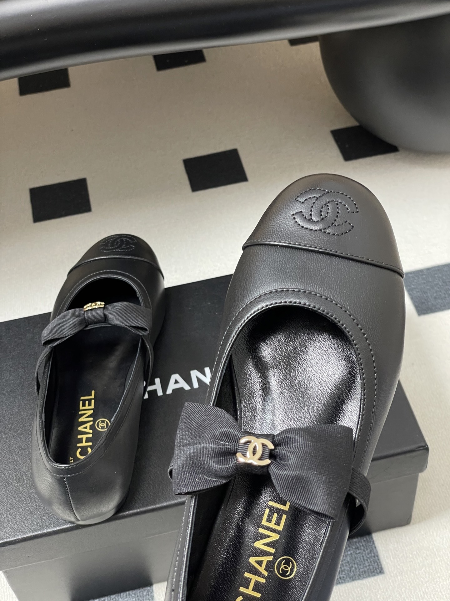 A pair of black Chanel leather ballerina flats featuring a grosgrain bow and gold-tone CC charm on a black table. - Hình 9