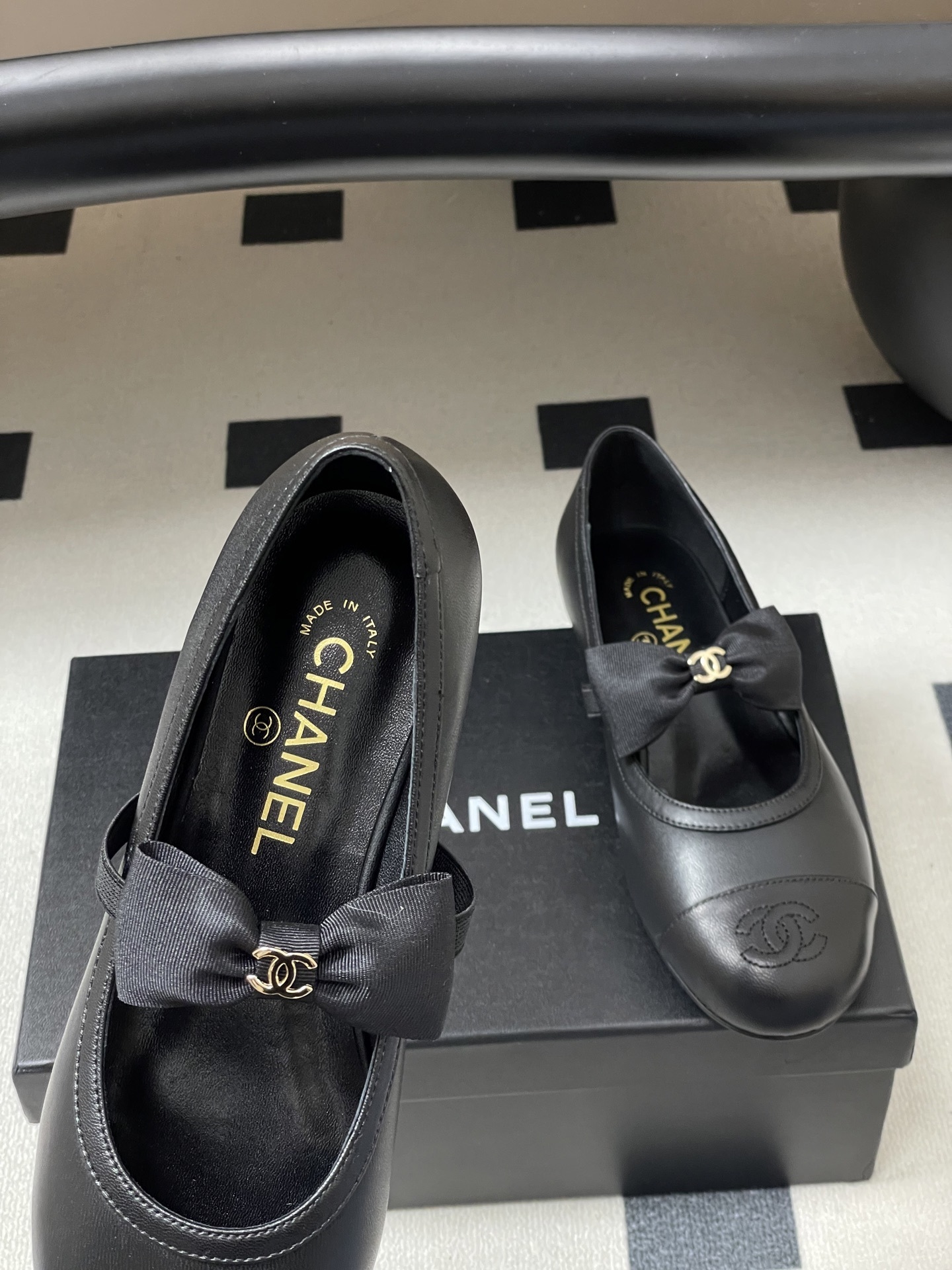 A pair of black Chanel leather ballerina flats featuring a grosgrain bow and gold-tone CC charm on a black table. - Hình 7