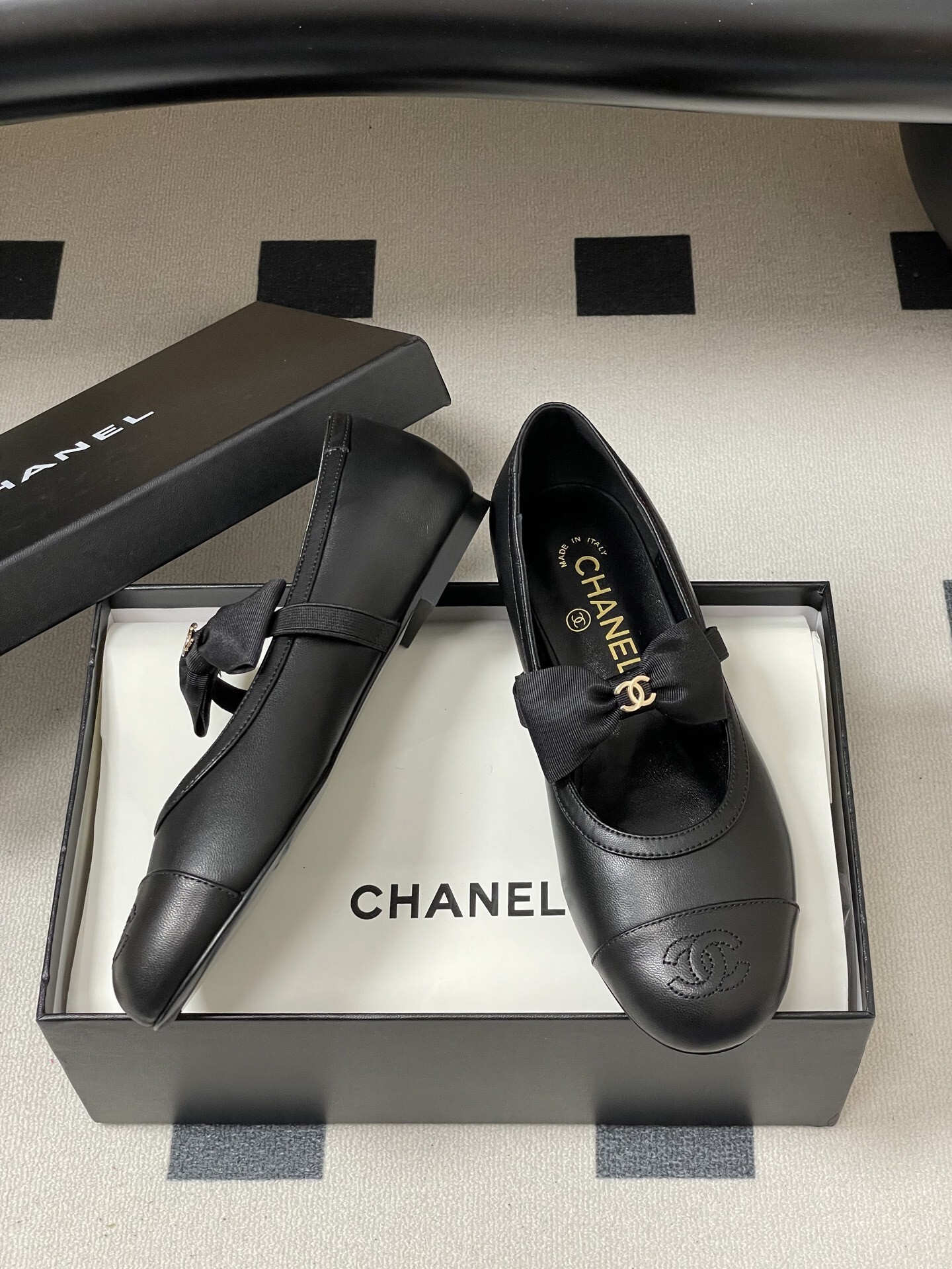A pair of black Chanel leather ballerina flats featuring a grosgrain bow and gold-tone CC charm on a black table. - Hình 6