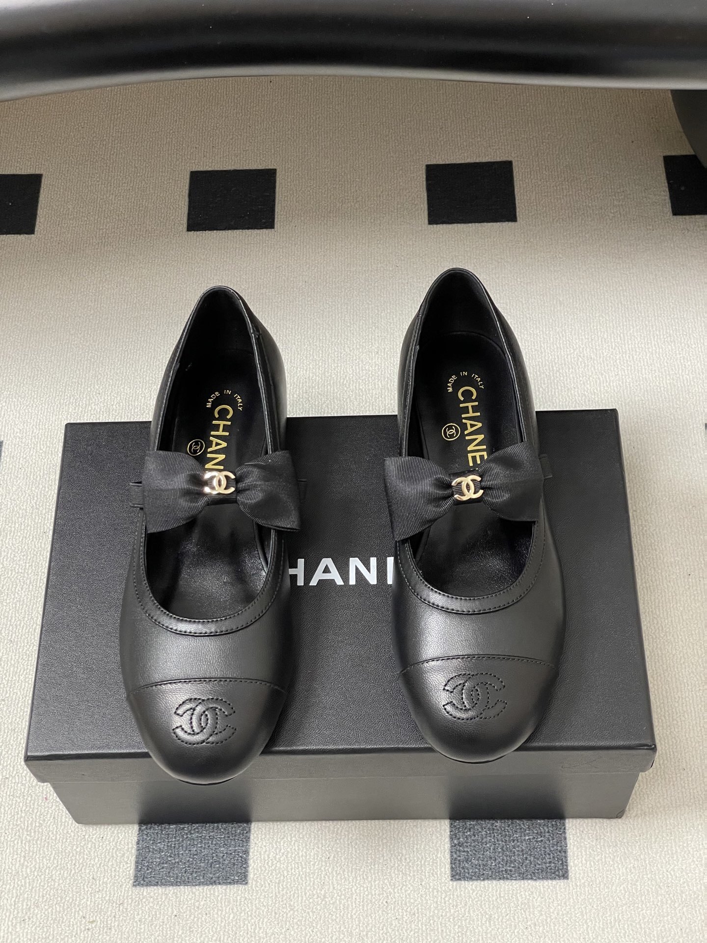 A pair of black Chanel leather ballerina flats featuring a grosgrain bow and gold-tone CC charm on a black table. - Hình 5