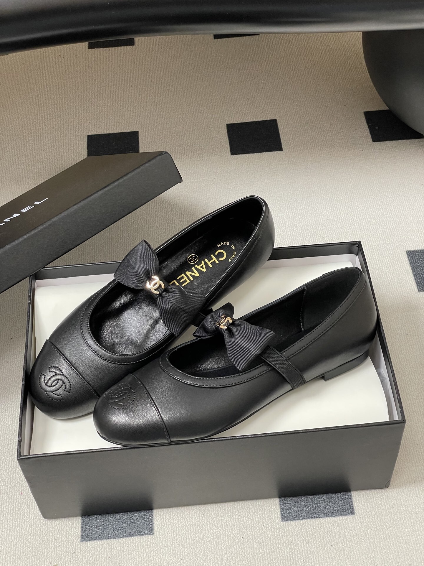 A pair of black Chanel leather ballerina flats featuring a grosgrain bow and gold-tone CC charm on a black table. - Hình 4