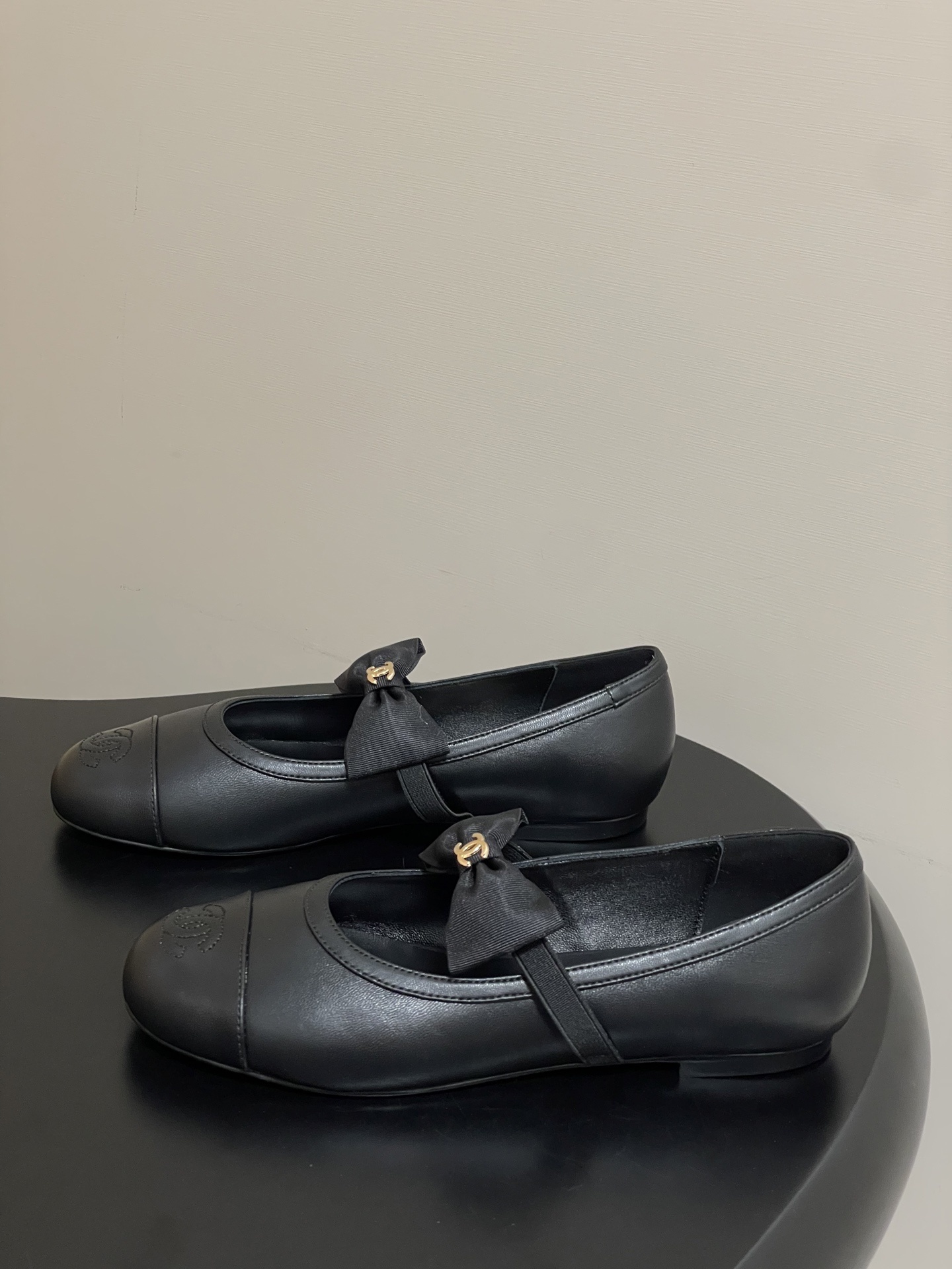 A pair of black Chanel leather ballerina flats featuring a grosgrain bow and gold-tone CC charm on a black table. - Hình 3