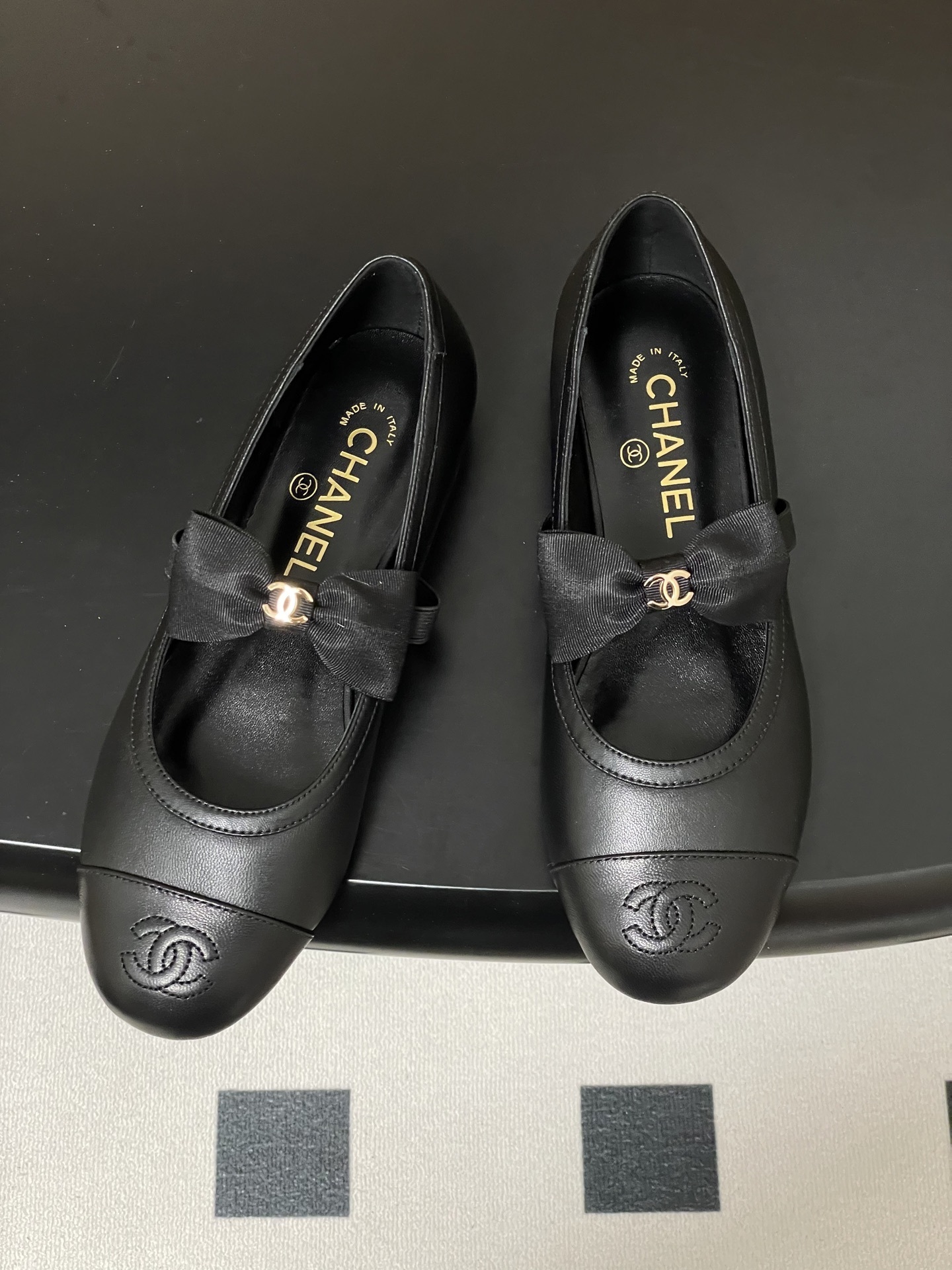 A pair of black Chanel leather ballerina flats featuring a grosgrain bow and gold-tone CC charm on a black table. - Hình 2