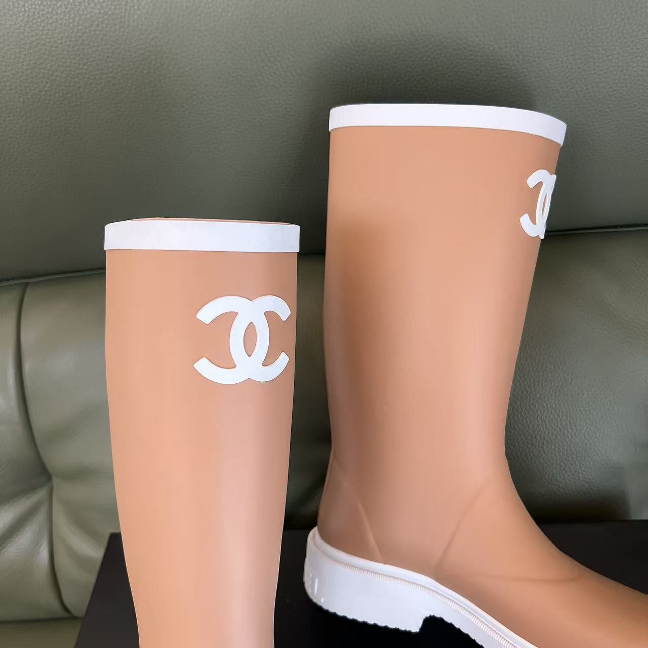 A pair of beige Chanel rain boots with white CC logo and white toe caps placed on a black designer box. - Hình 8
