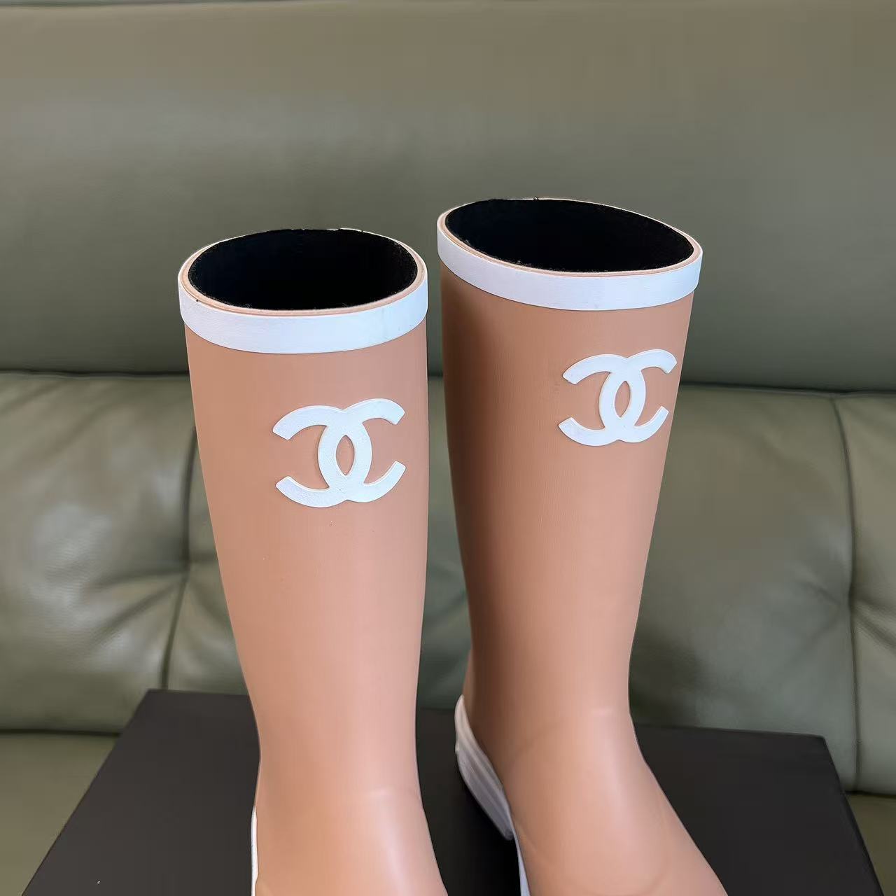A pair of beige Chanel rain boots with white CC logo and white toe caps placed on a black designer box. - Hình 5