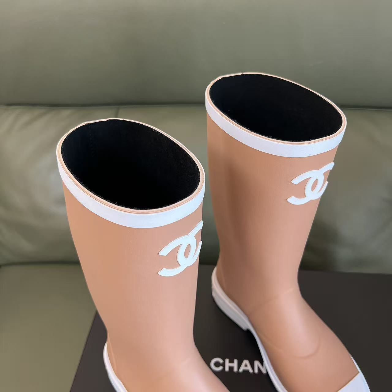 A pair of beige Chanel rain boots with white CC logo and white toe caps placed on a black designer box. - Hình 4