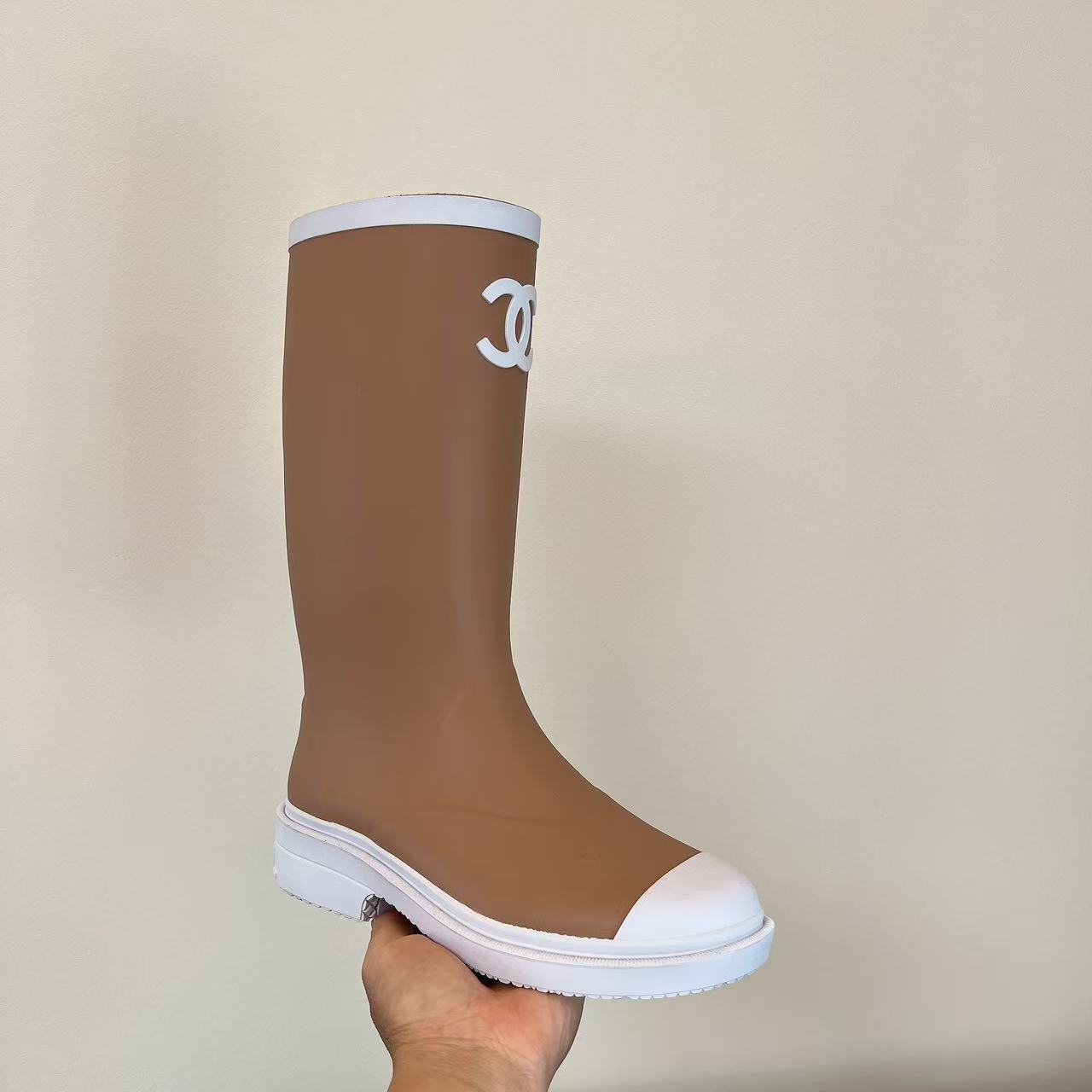 A pair of beige Chanel rain boots with white CC logo and white toe caps placed on a black designer box. - Hình 3