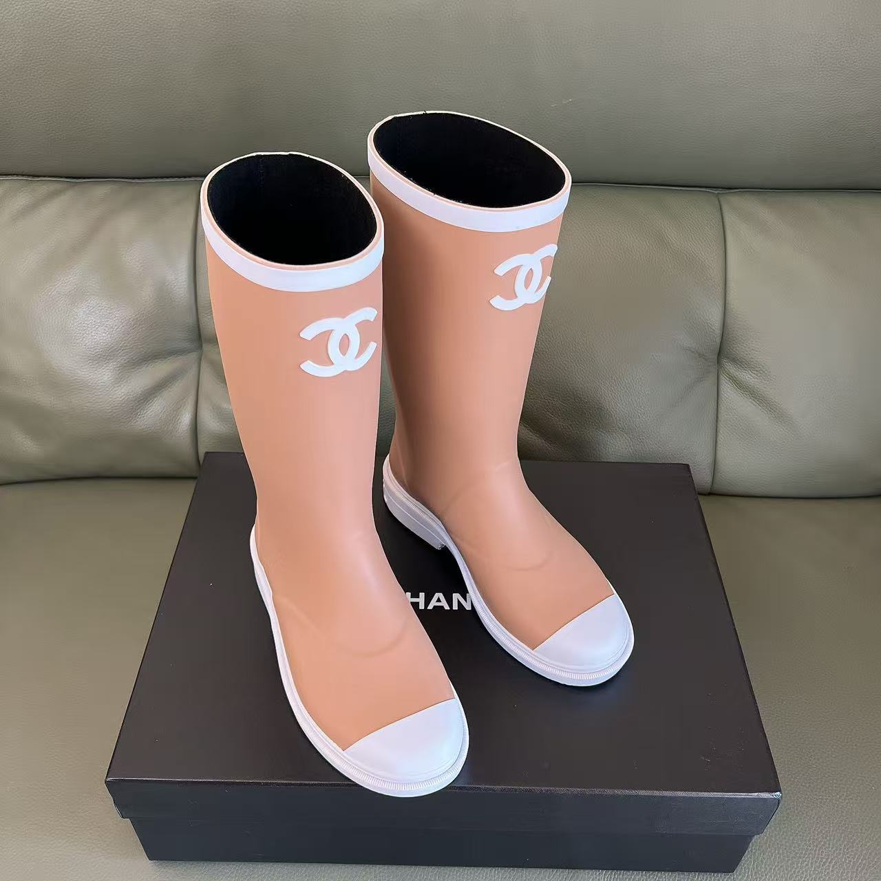 A pair of beige Chanel rain boots with white CC logo and white toe caps placed on a black designer box. - Hình 2