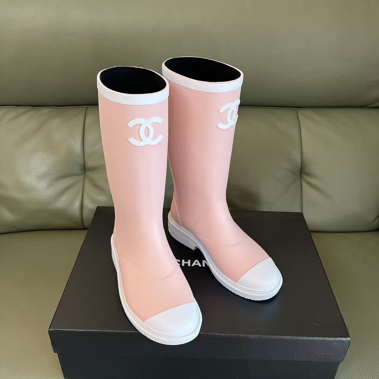 A pair of light pink luxury rubber rain boots with a white interlocking logo and white toe caps placed on a black shoe box. - Hình 2